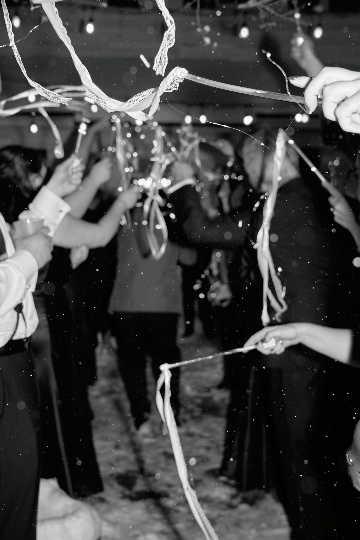Black and white photo of wedding guests holding sparklers and ribbons during a festive outdoor celebration.