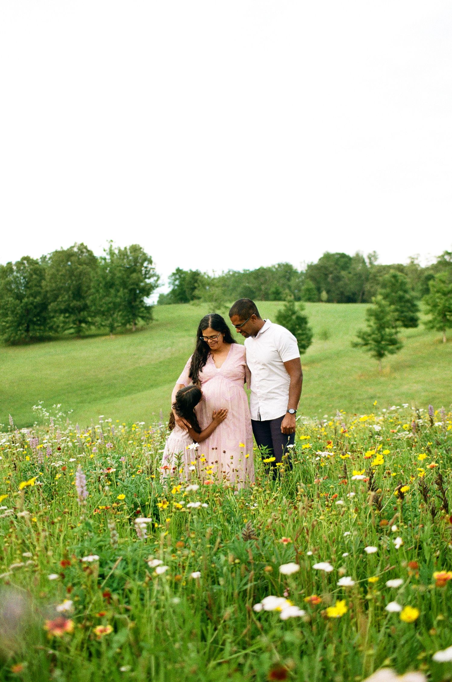 Pregnant couple standing in a wildflower meadow, woman in pink dress, man in white shirt, lush green hills behind them.