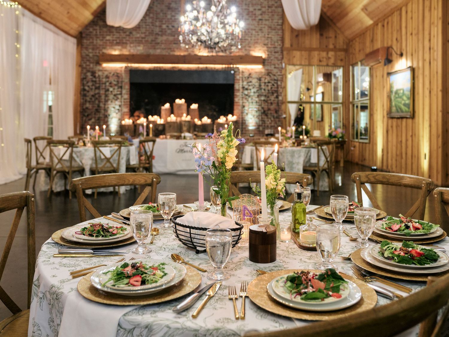 Elegant barn wedding reception with round tables, floral centerpieces, gold chargers, and chandelier lighting in a rustic venue.