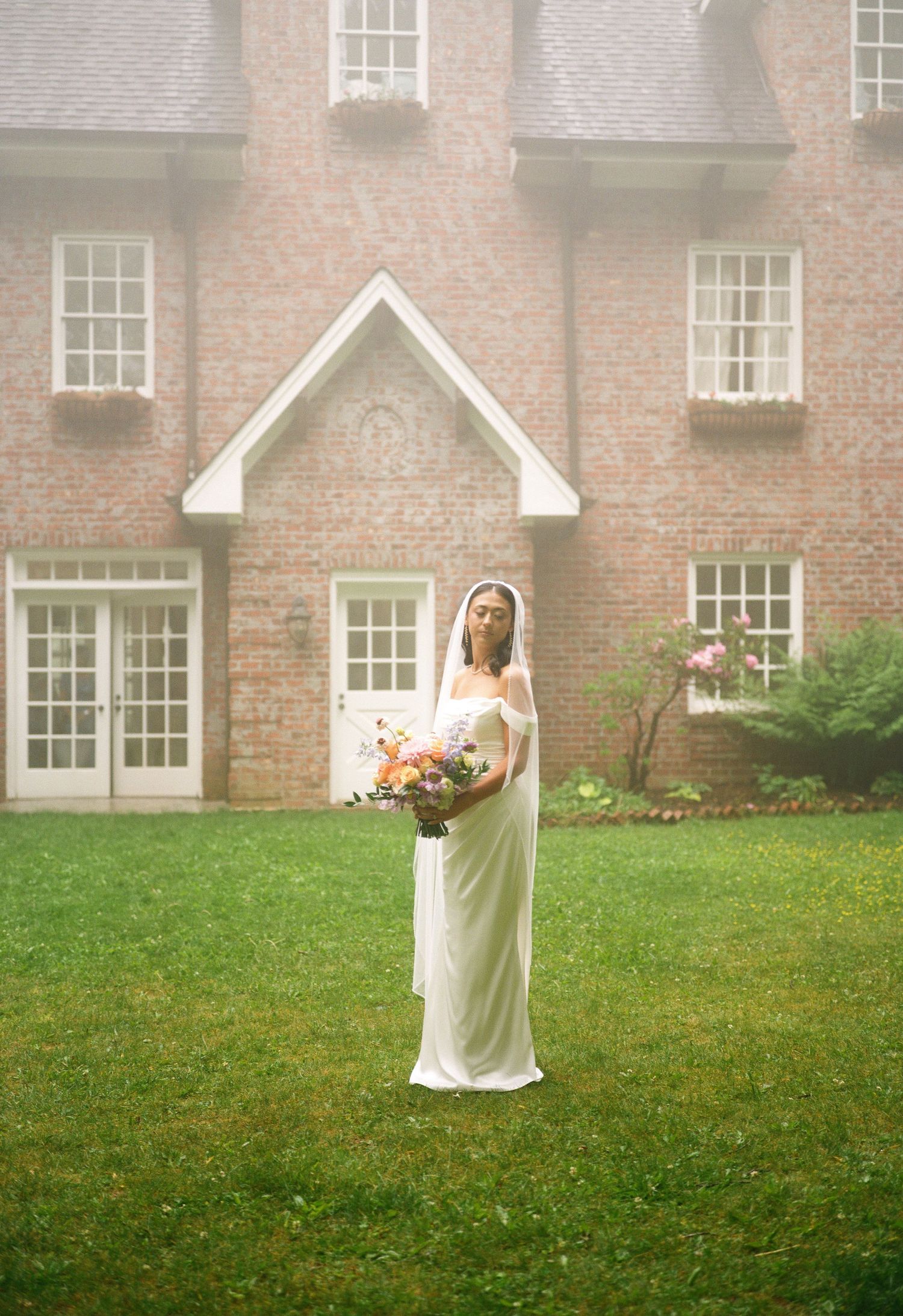 Bride in white gown and veil holds colorful bouquet on green lawn before elegant brick manor.