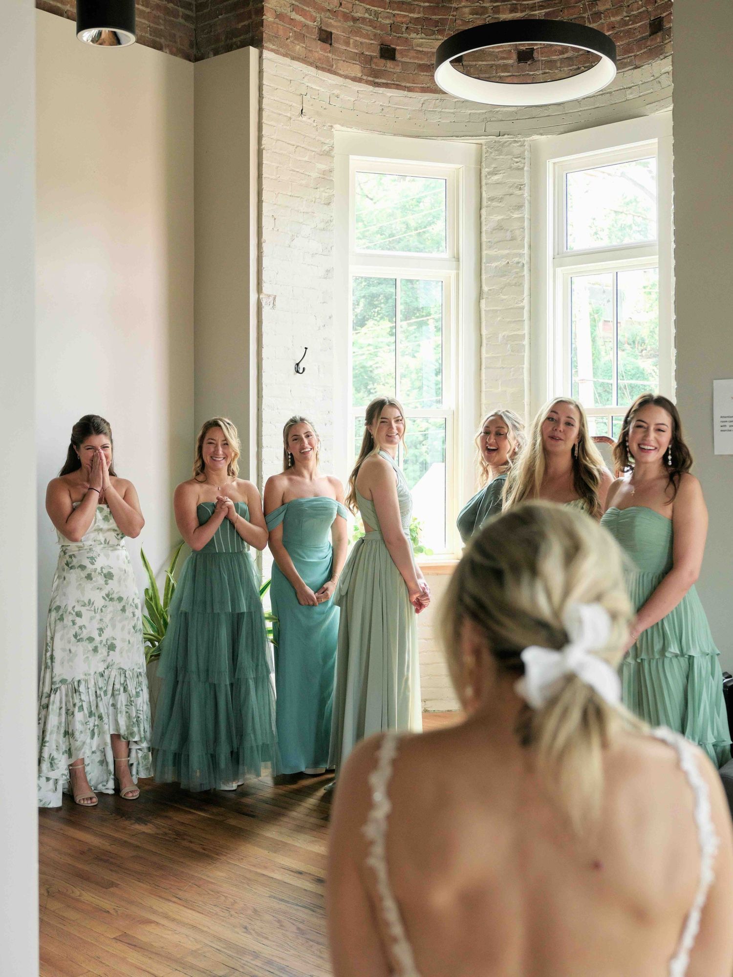 Bridesmaids in sage green dresses react emotionally as bride reveals her wedding gown in a bright, elegant bridal suite.