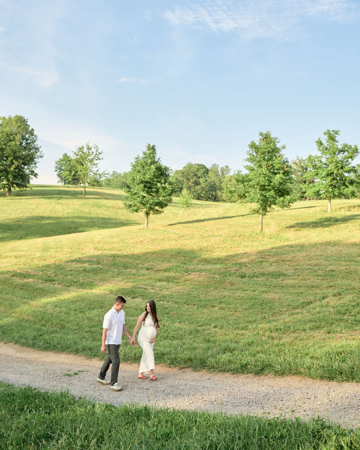 A couple walks hand-in-hand on a gravel path surrounded by lush green rolling hills and trees under a bright blue sky.