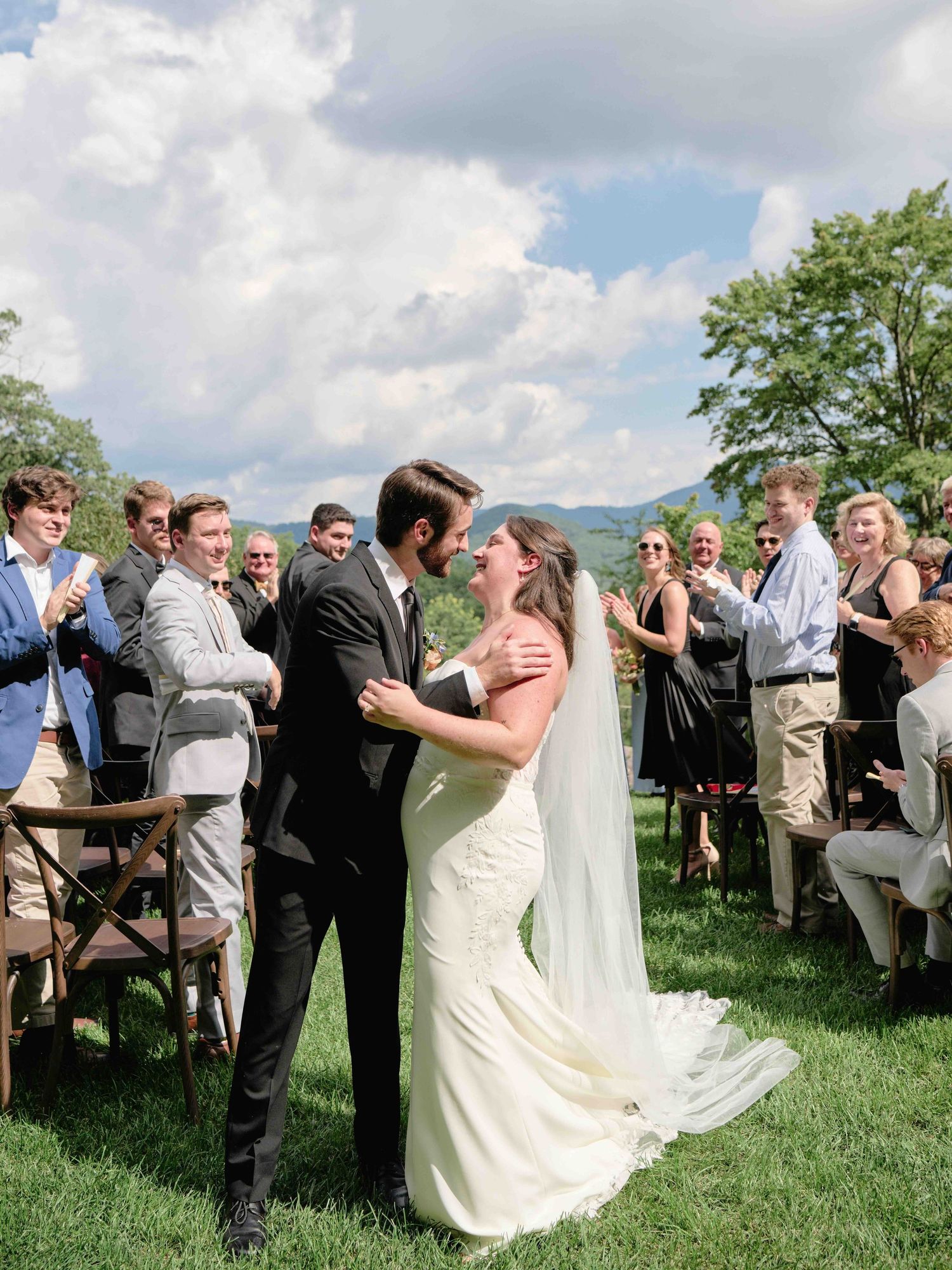 Bride and groom share first kiss at outdoor wedding ceremony with mountain backdrop and cheering guests.