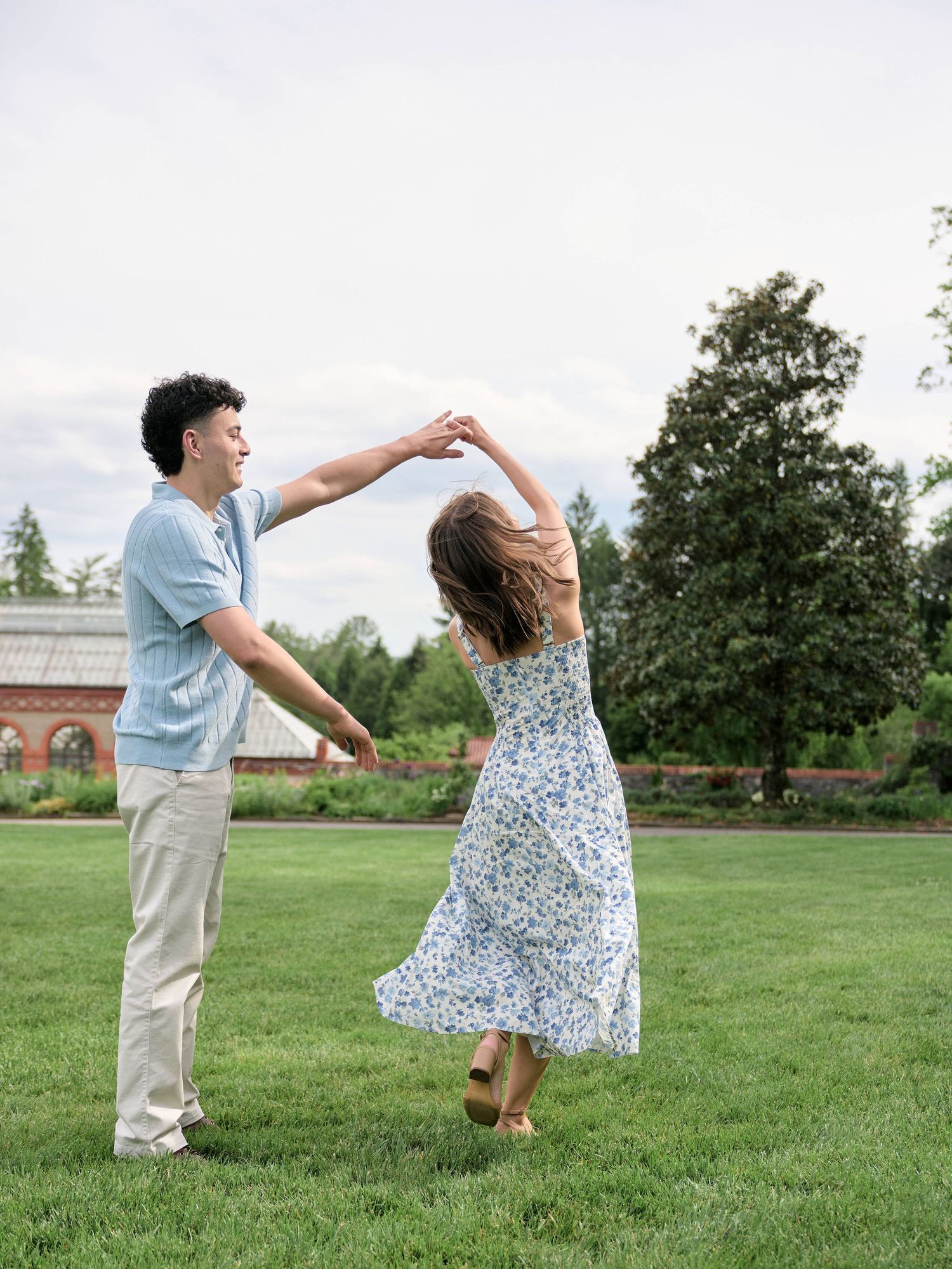 A couple dances joyfully on a green lawn, the woman in a floral dress spinning as the man holds her hand up.