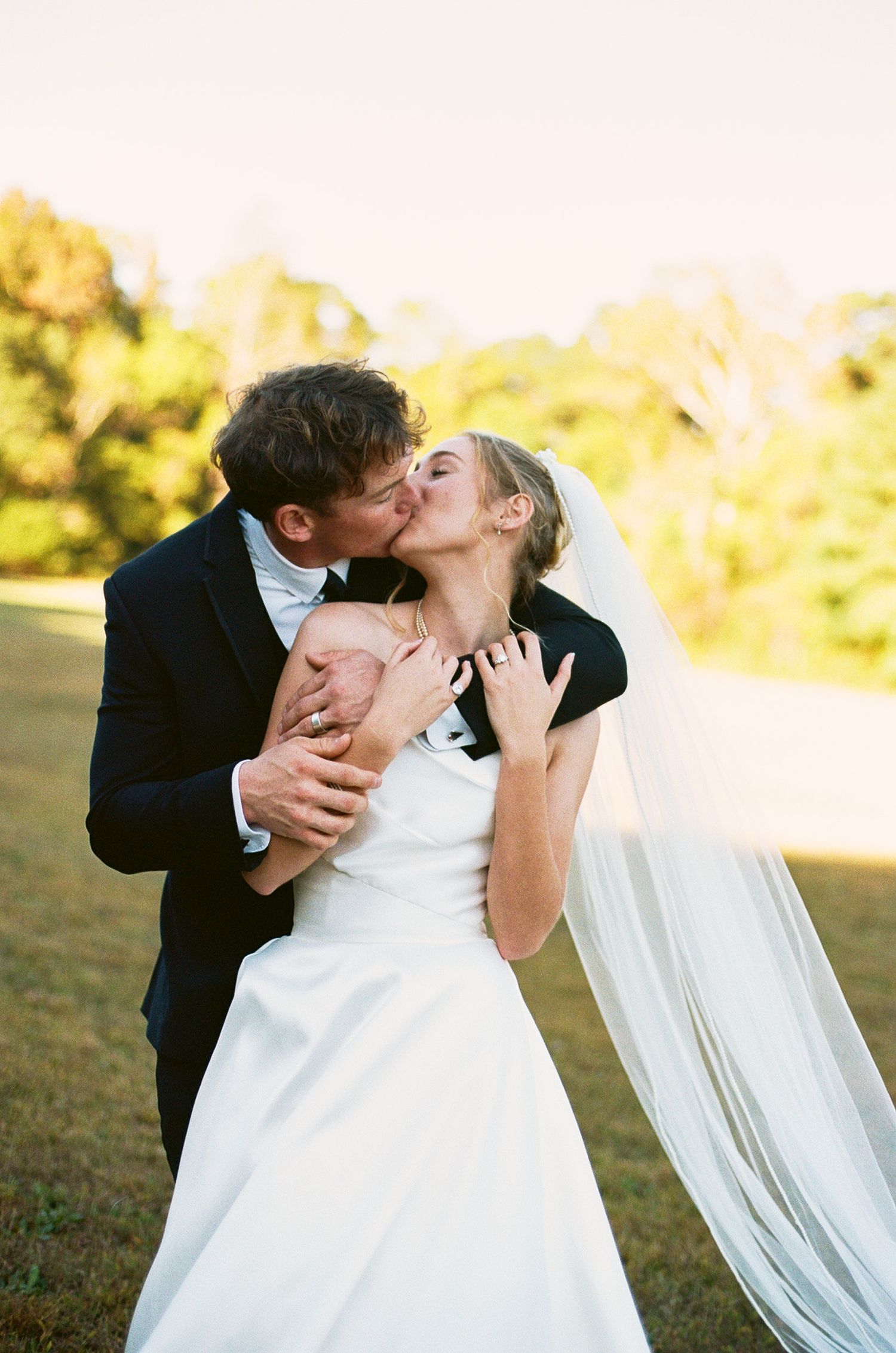 A romantic wedding couple shares a passionate dip kiss in a sunlit outdoor setting.