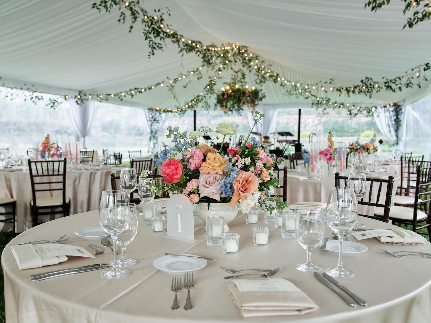 Elegant wedding reception table setting under white tent with floral centerpiece and twinkling string lights overhead.