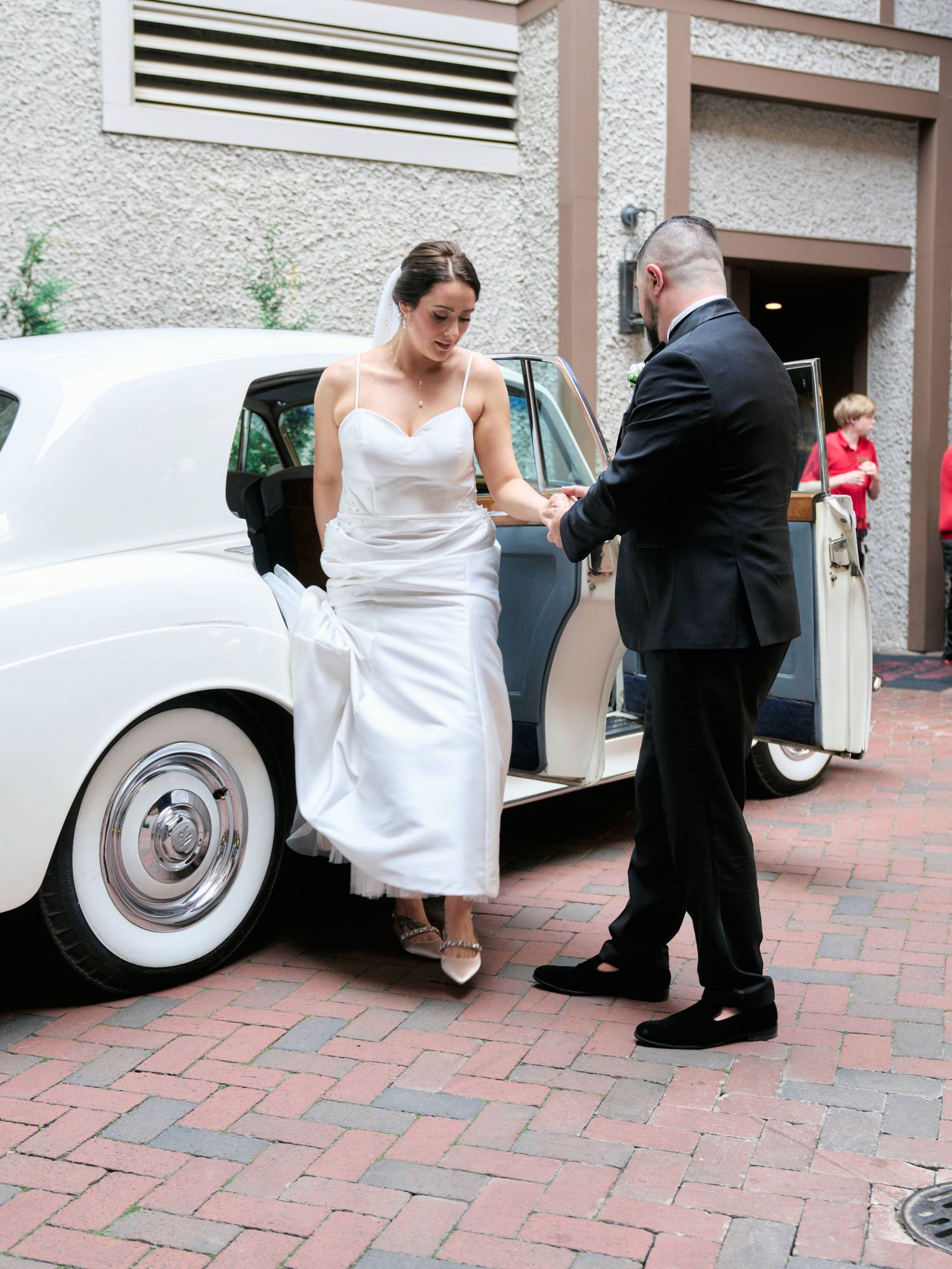 A bride in a white wedding dress steps out of a classic vintage car with assistance on a brick paved driveway.