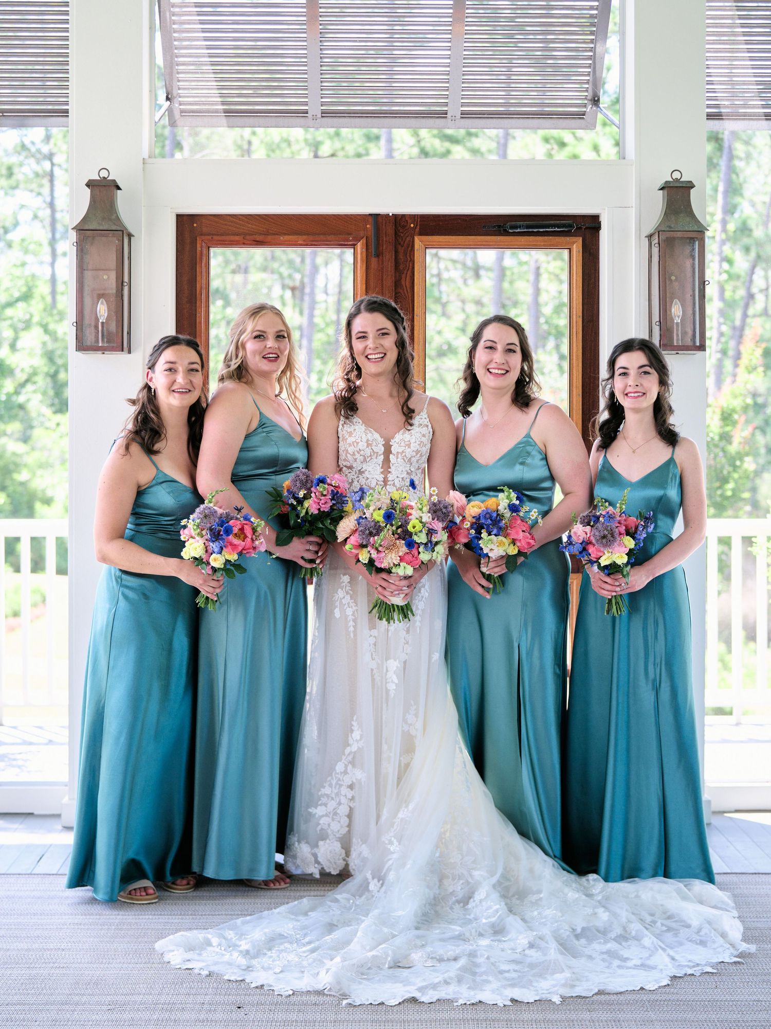 A bridal party in teal bridesmaid dresses and a white lace wedding gown pose on a covered porch with lanterns.