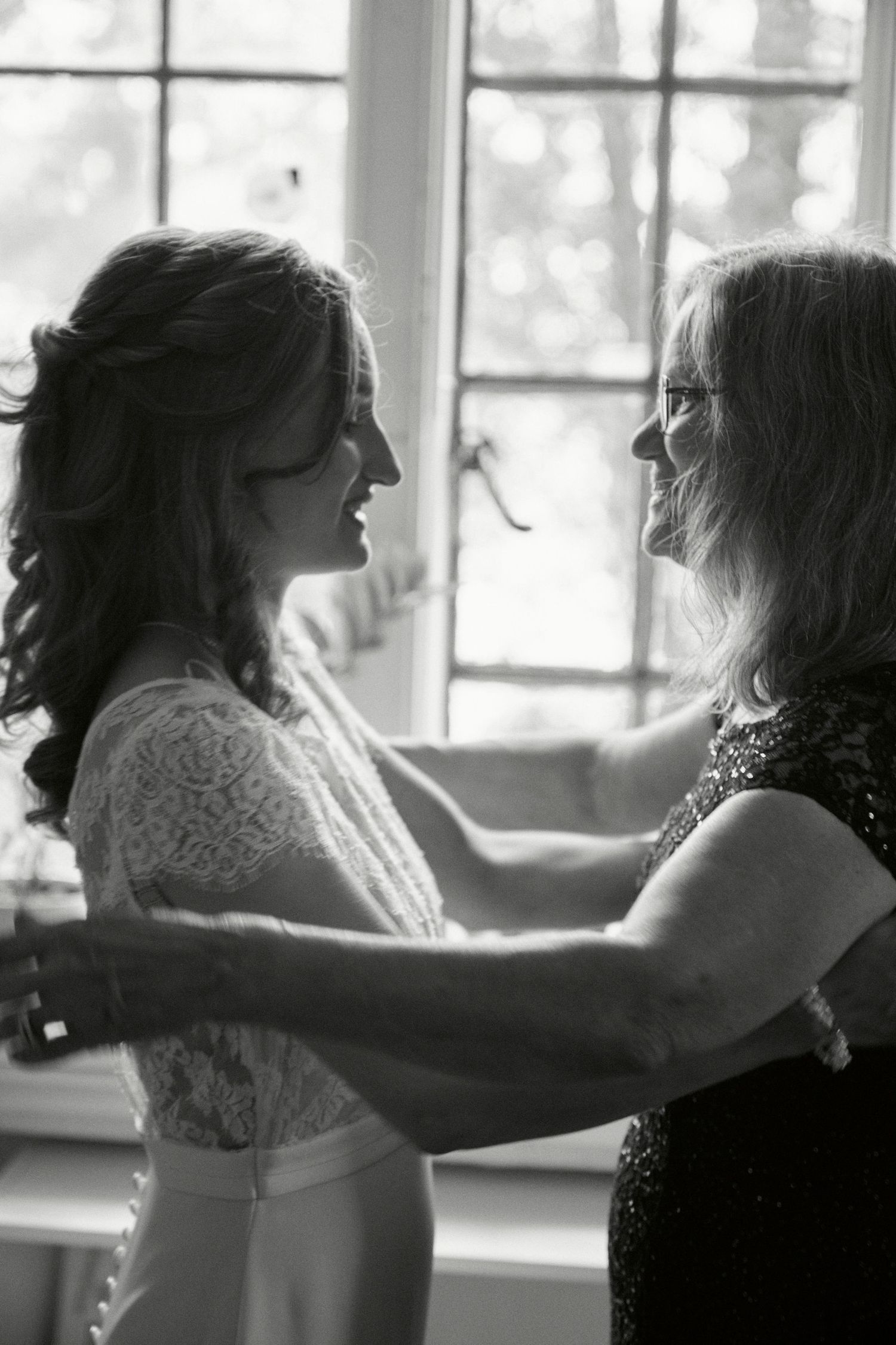 Black and white candid moment between two women by window in wedding attire.