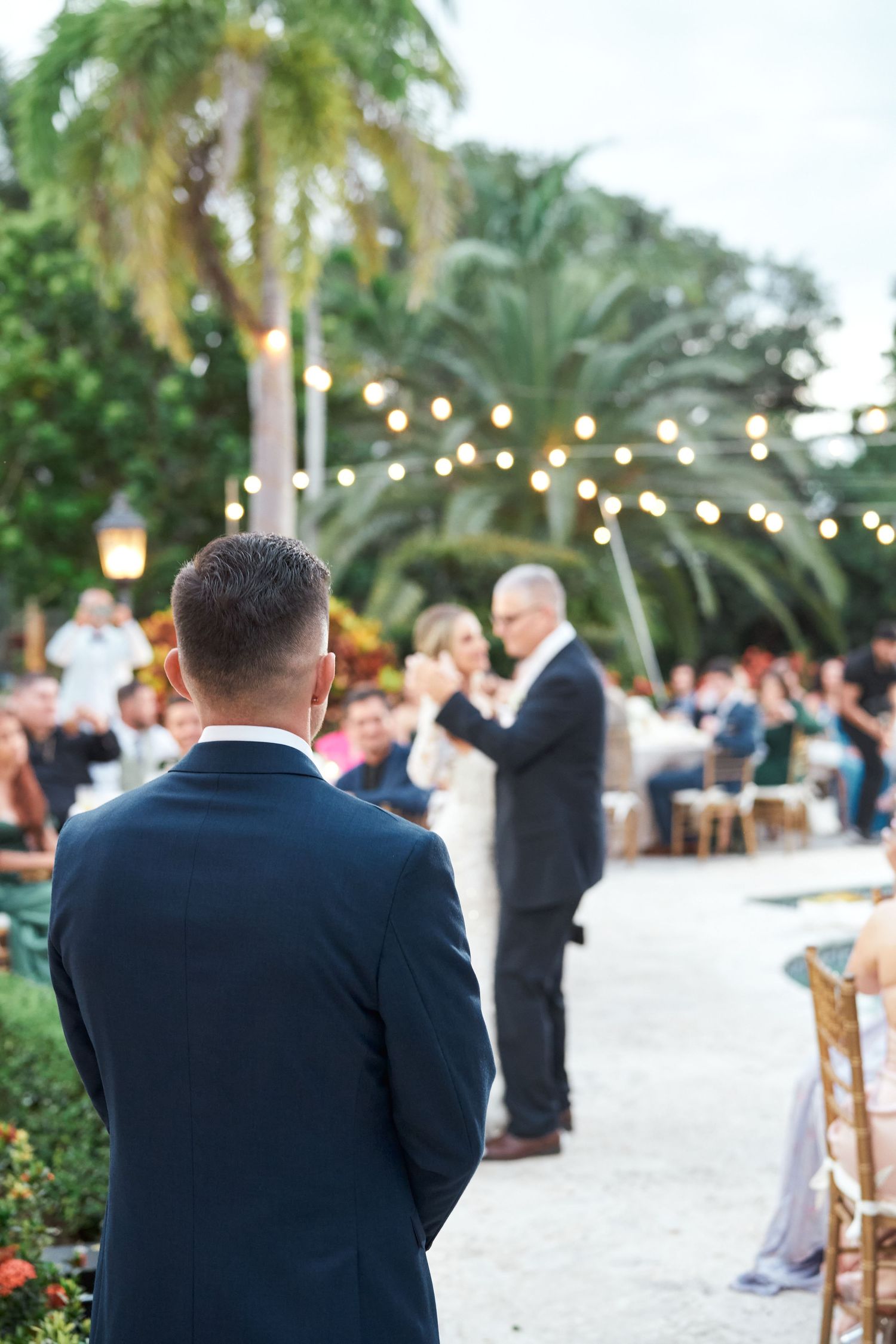 Navy-suited guest watches outdoor wedding ceremony under string lights and palm trees.