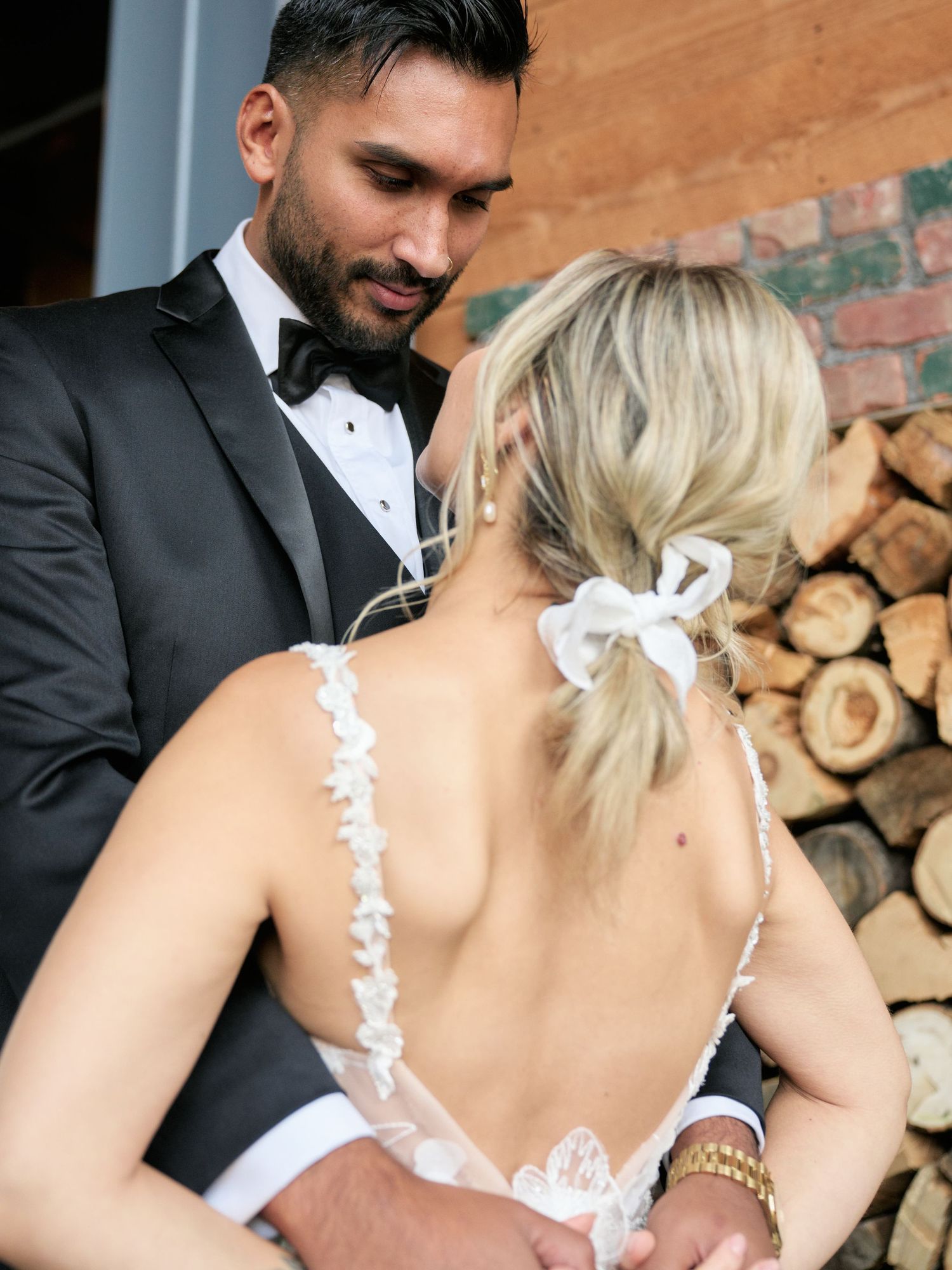 A bride and groom share an intimate moment by a rustic wood pile backdrop, her backless lace dress elegantly detailed.
