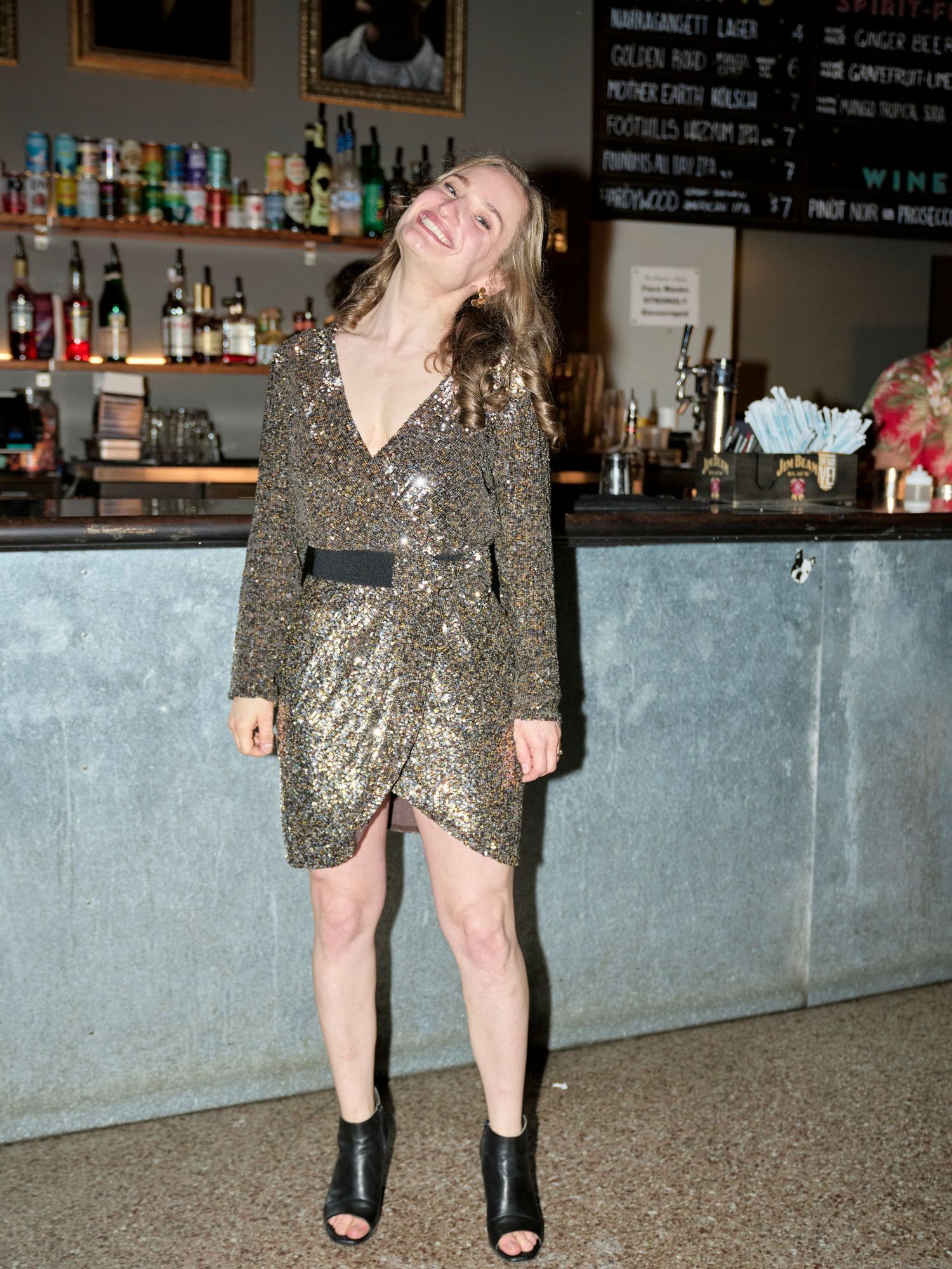 Woman in a sparkly dress poses playfully in front of a bar counter.