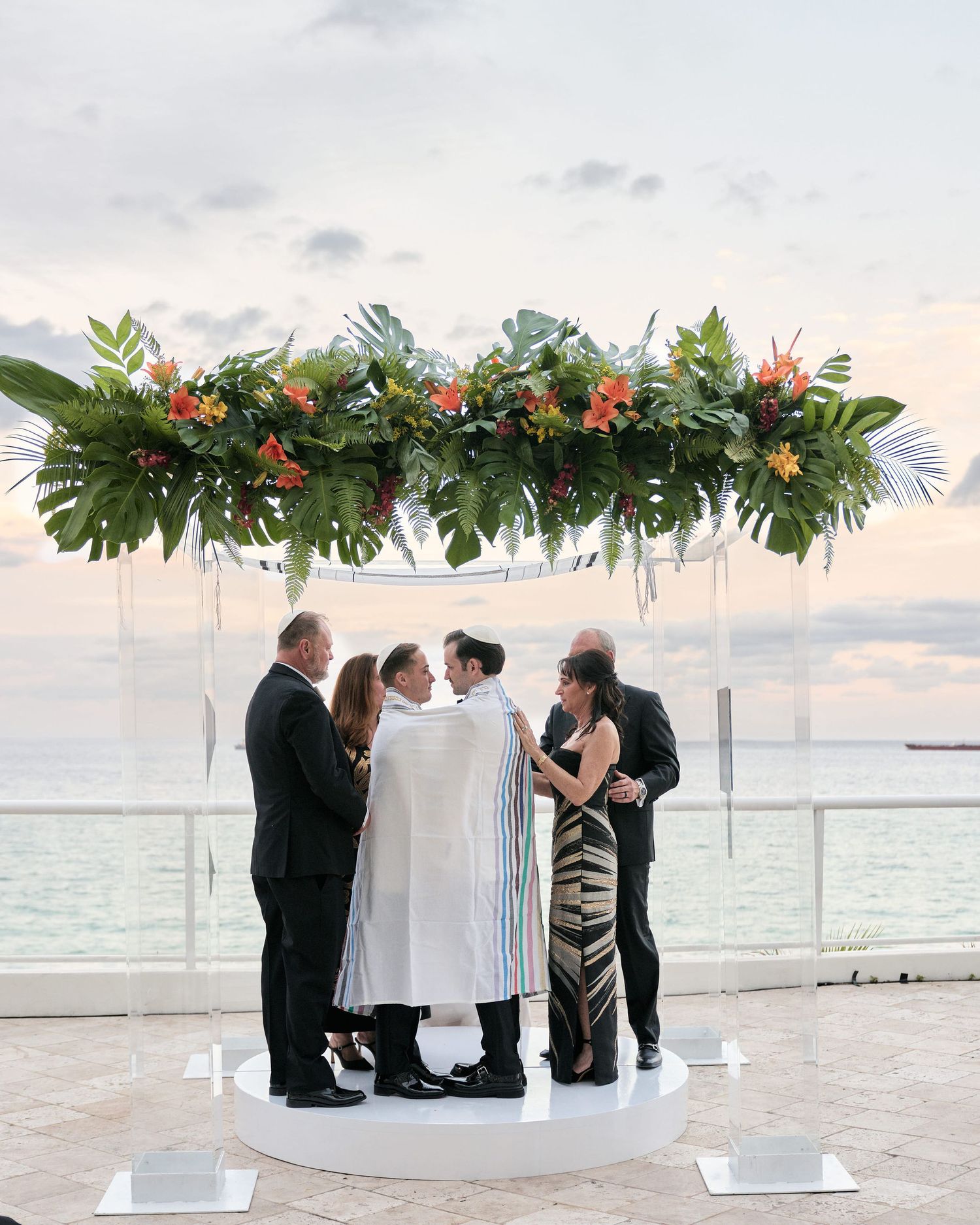A wedding ceremony takes place under a tropical floral arch overlooking the ocean at sunset.