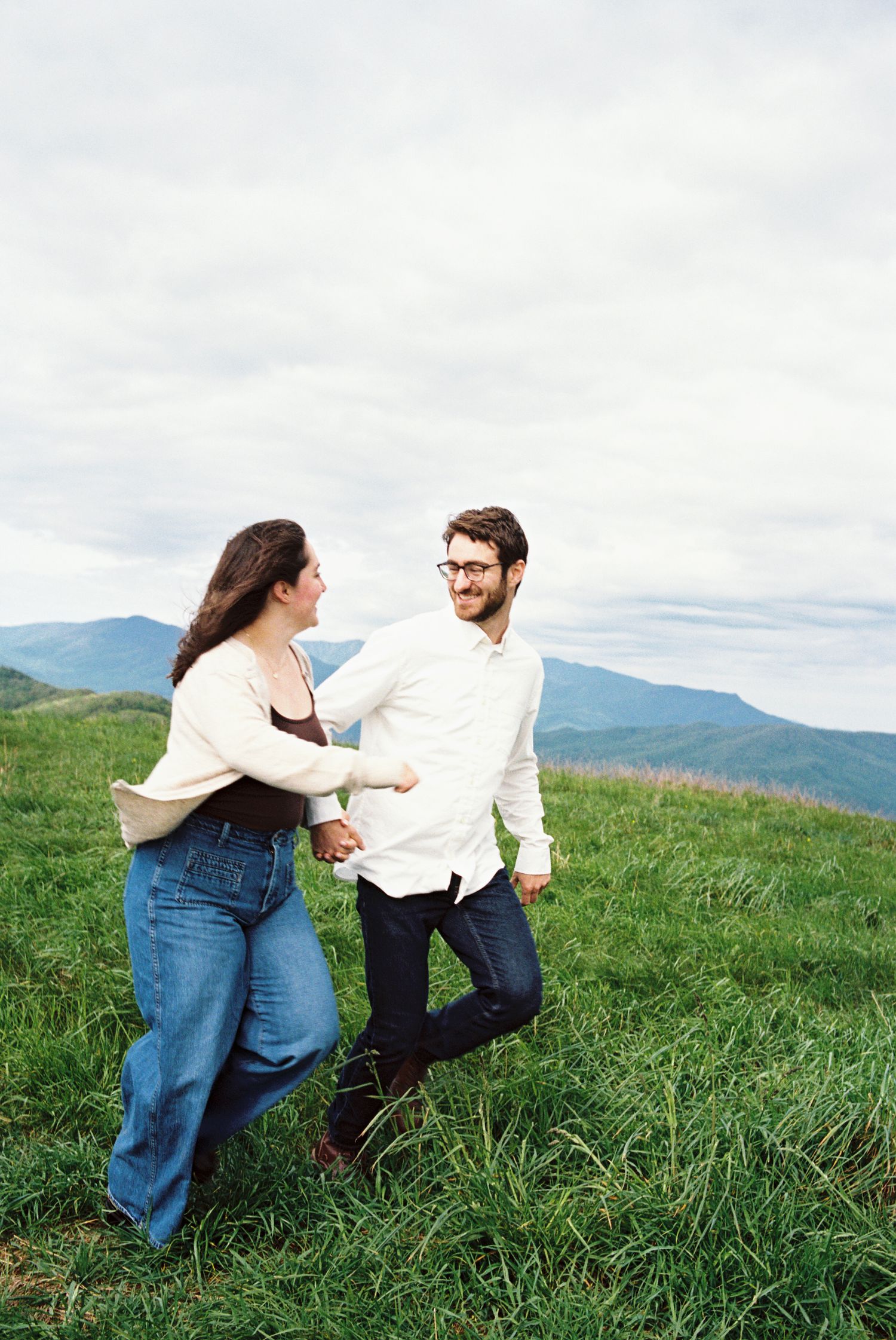A couple enjoying a playful moment together on a grassy hillside with mountain views in the background.