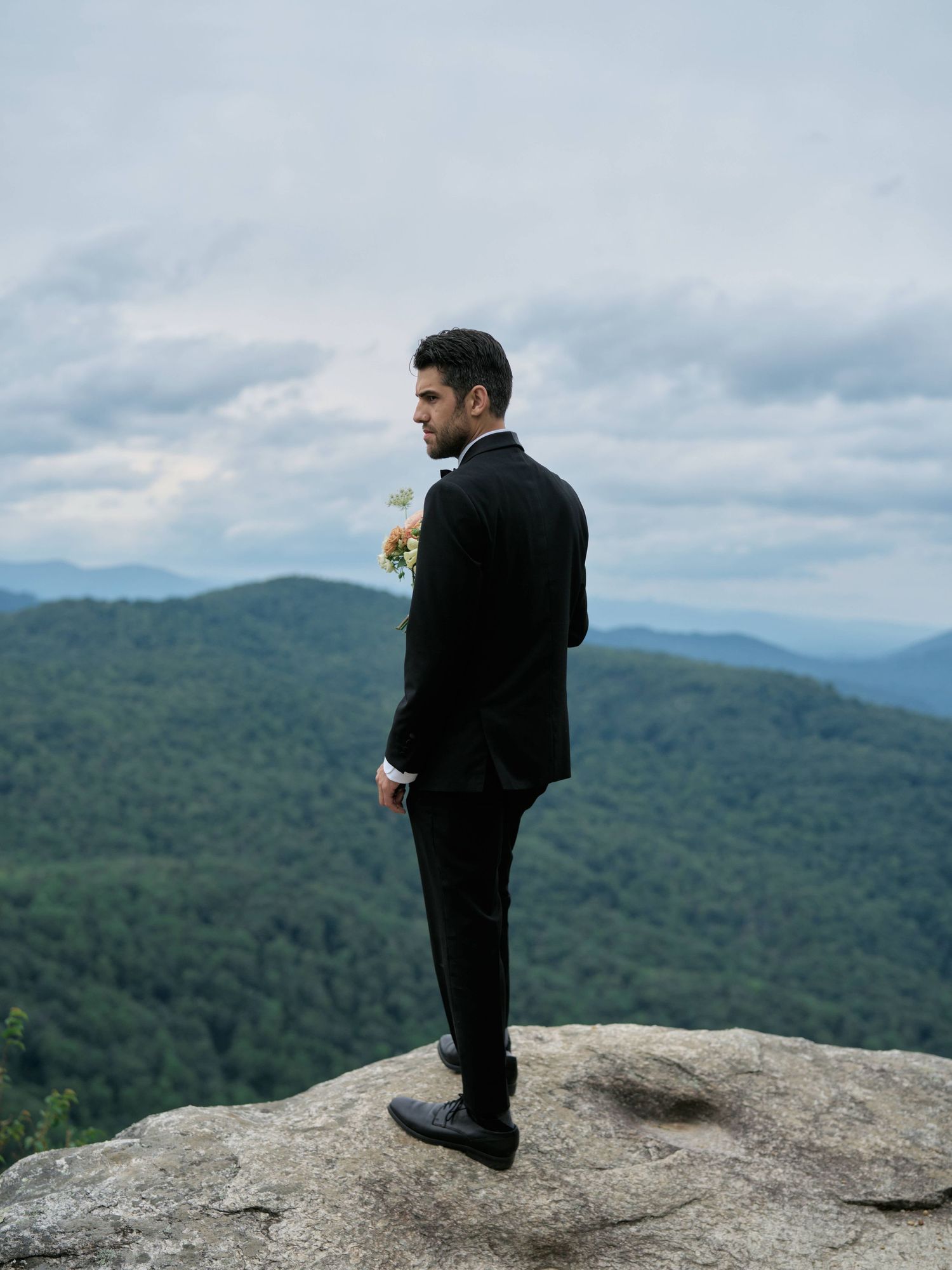 A person in black clothing stands on a rocky mountain overlook with sweeping views of forested peaks and cloudy skies.