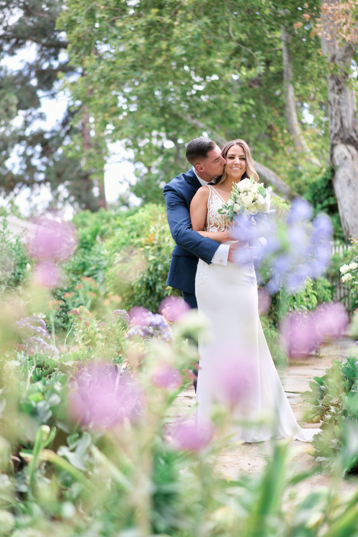 Dreamy garden wedding photo with couple embracing among purple flowers.