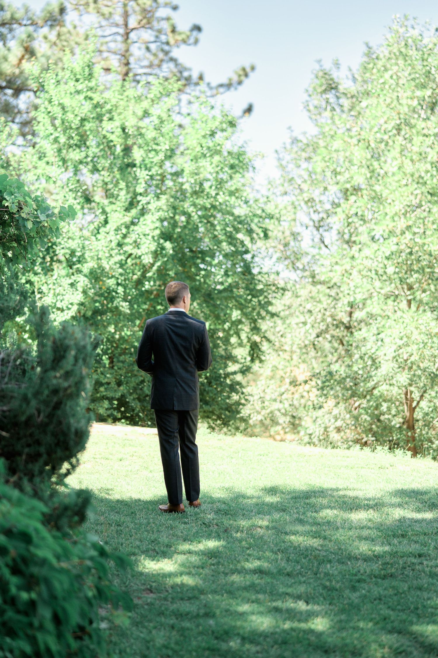 Person in black suit standing alone in a lush green garden setting.