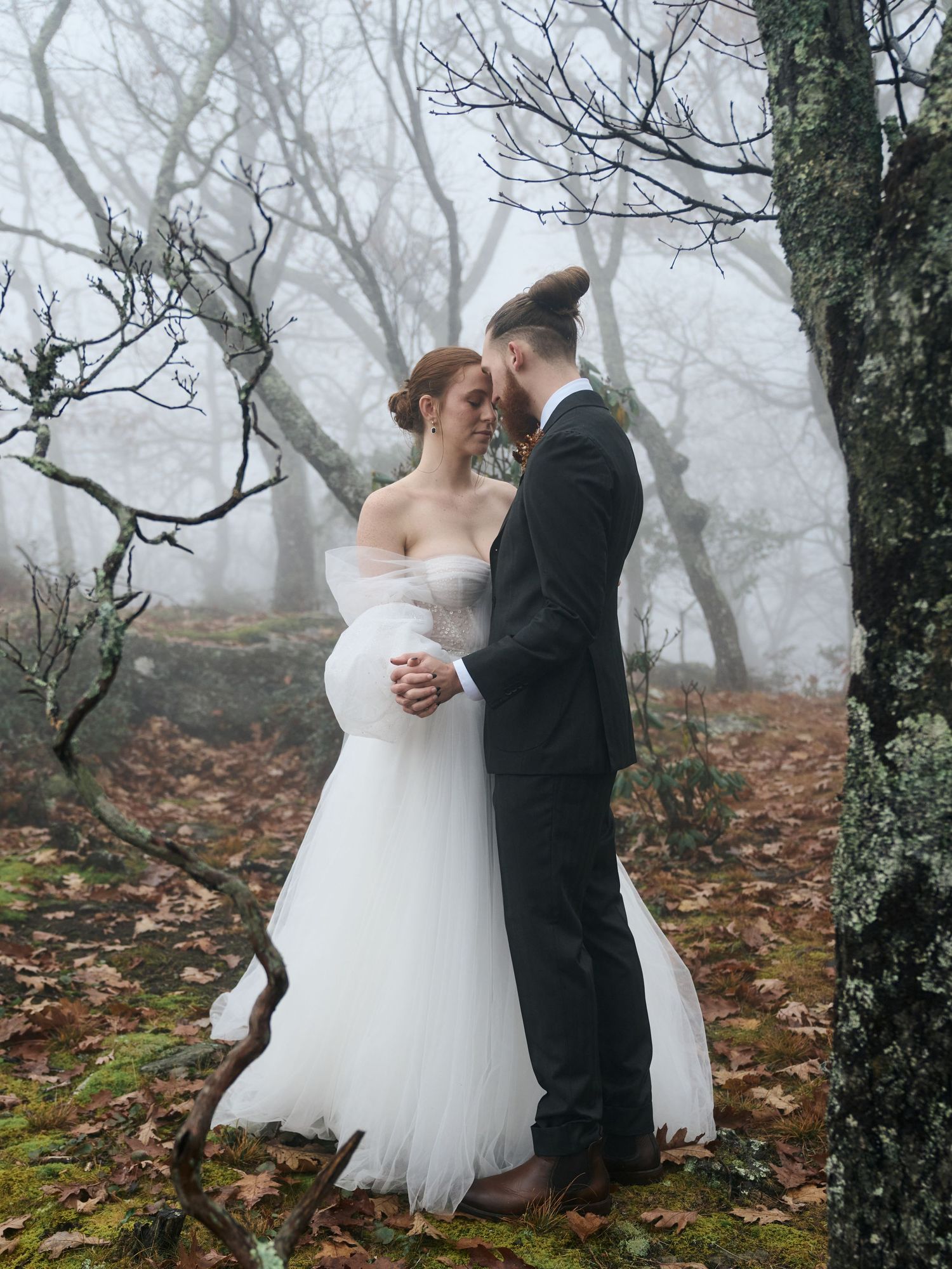 A tender moment between newlyweds in a misty forest with bare trees and fallen leaves.