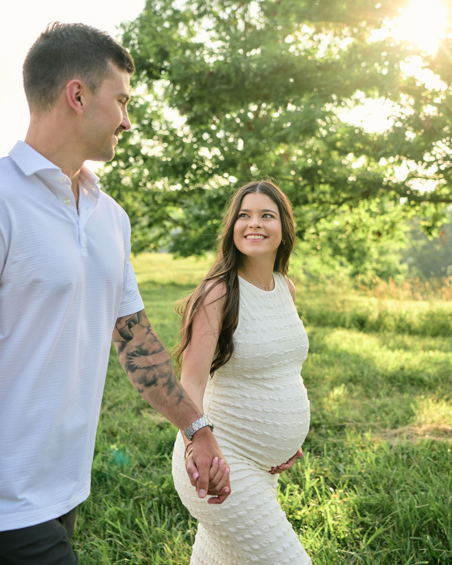 Pregnant woman in white dress holds hands with tattooed man outdoors in sunny green field during maternity photo shoot.
