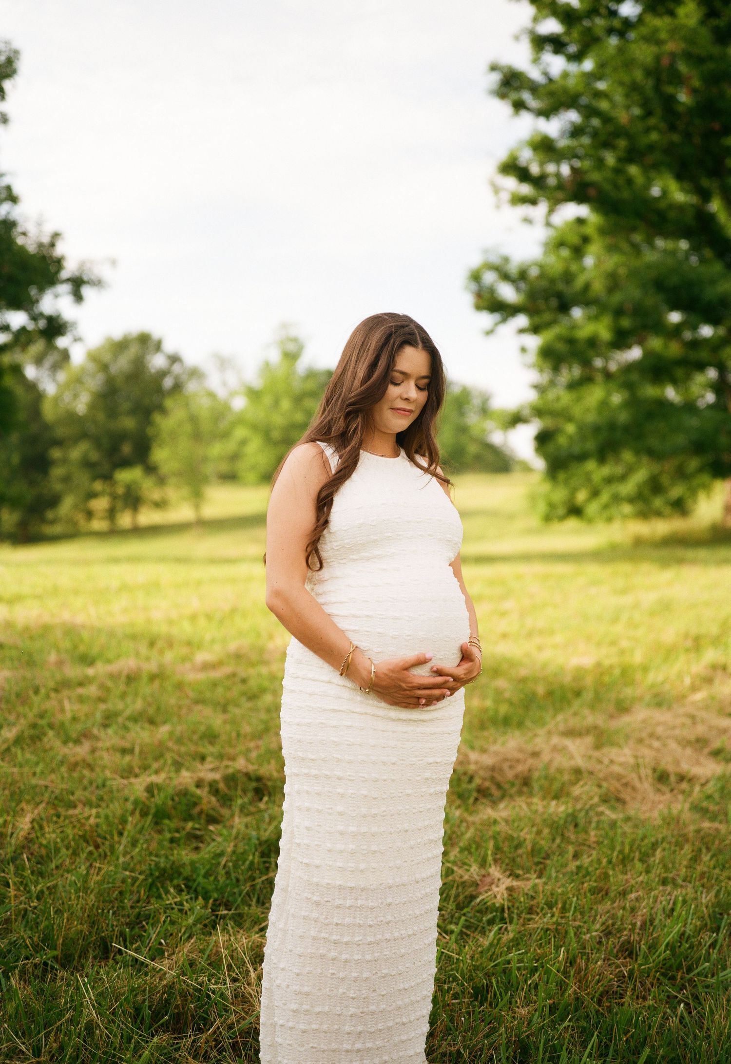 Pregnant woman in white dress cradling baby bump while standing in a lush green field during maternity shoot.