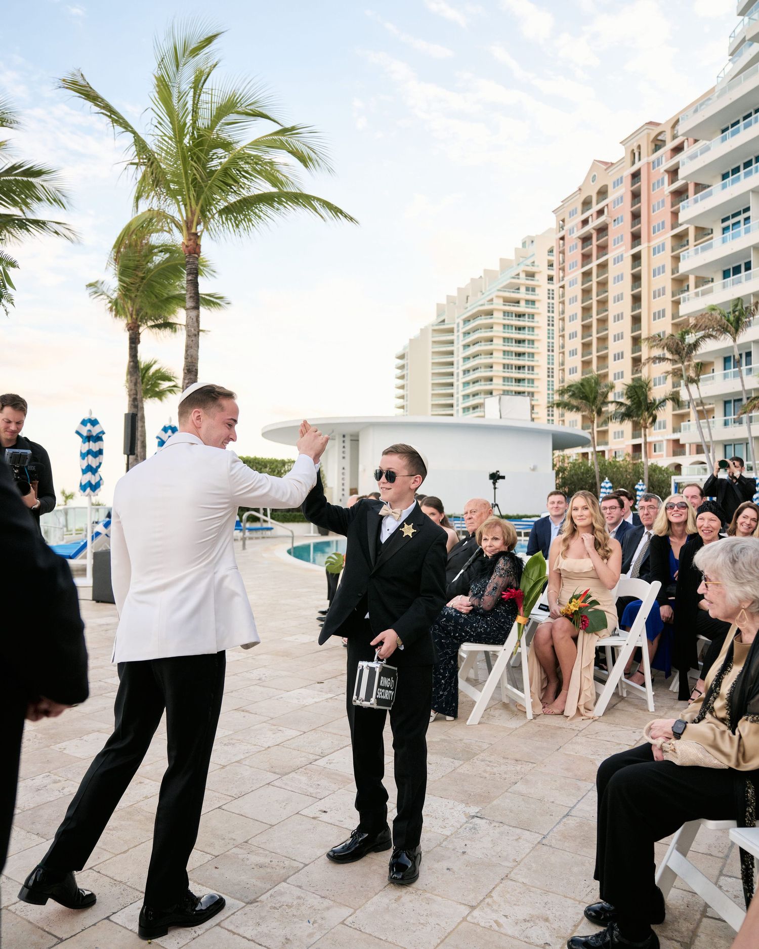 Wedding party performs a synchronized dance move on a beachfront patio with palm trees and condos in the background.