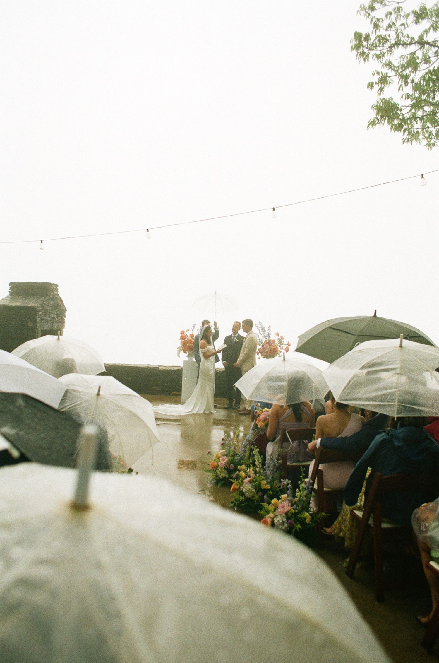 Rainy outdoor wedding ceremony with guests holding clear umbrellas as couple exchanges vows on a misty terrace.