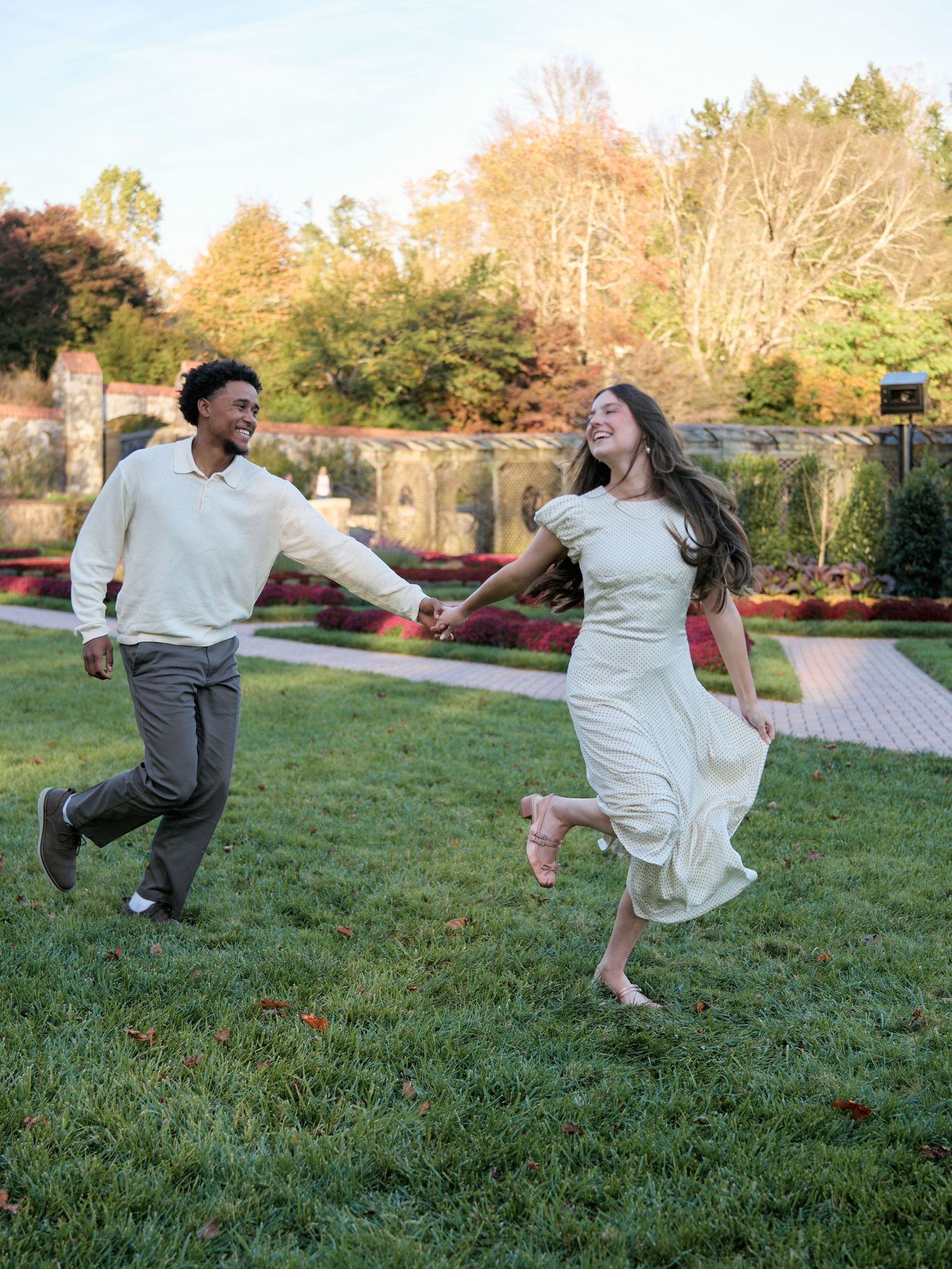 A couple dances joyfully on a manicured lawn with autumn trees and stone architecture in the background.