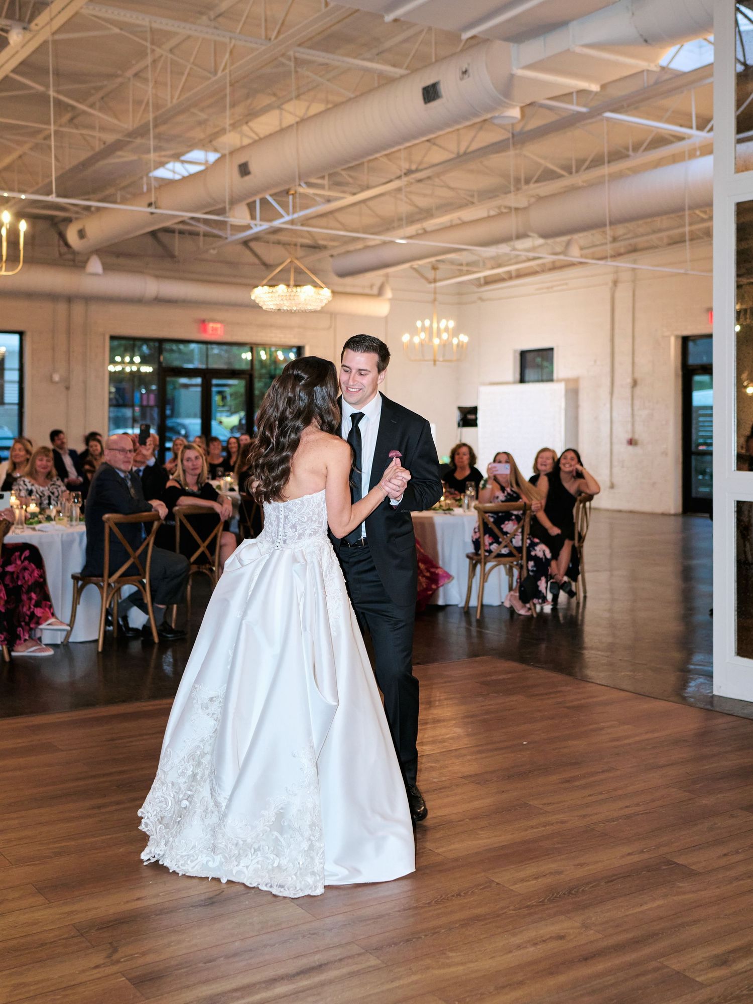 Bride and groom share their first dance at elegant wedding reception with wood floors and guests seated at candlelit tables.
