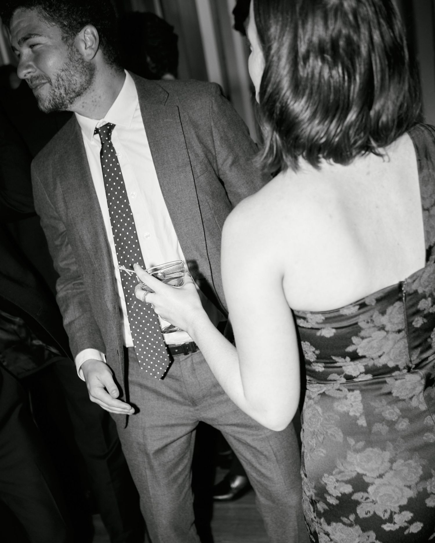 Black and white photo of a couple dancing at a formal event, man in suit with polka dot tie holding a drink.