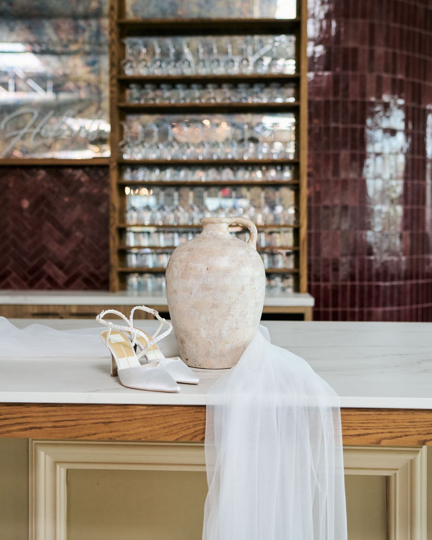 White bridal heels and rustic ceramic vase on marble counter with sheer veil, elegant bar shelving in background.