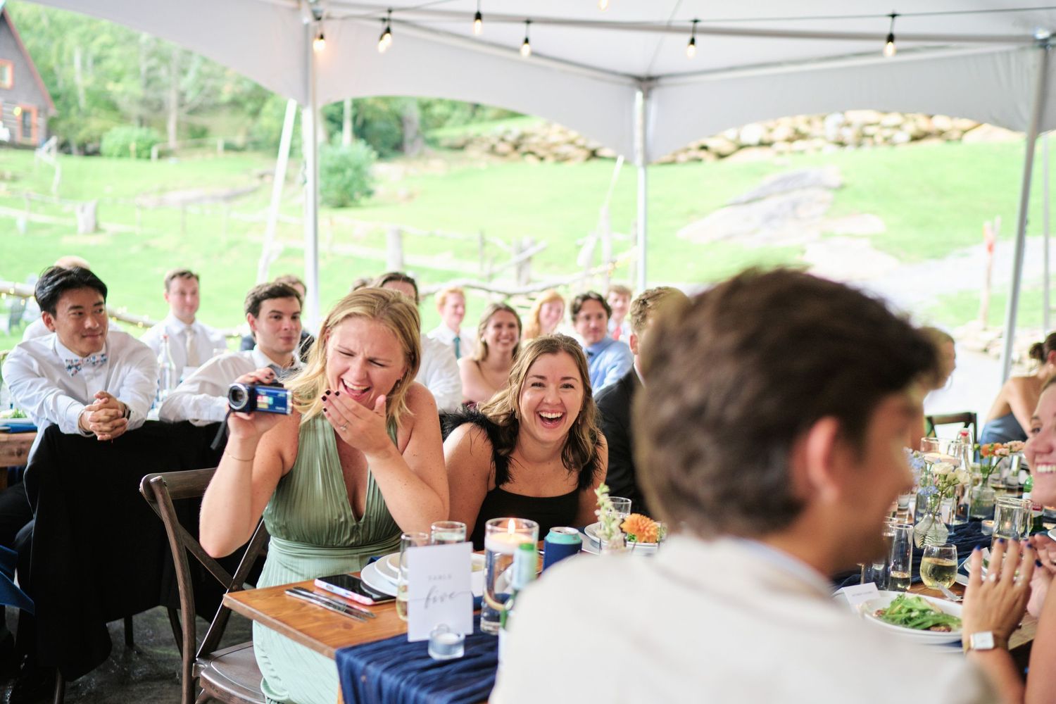 Guests laughing and celebrating at an outdoor wedding reception under a white tent on a sunny day.