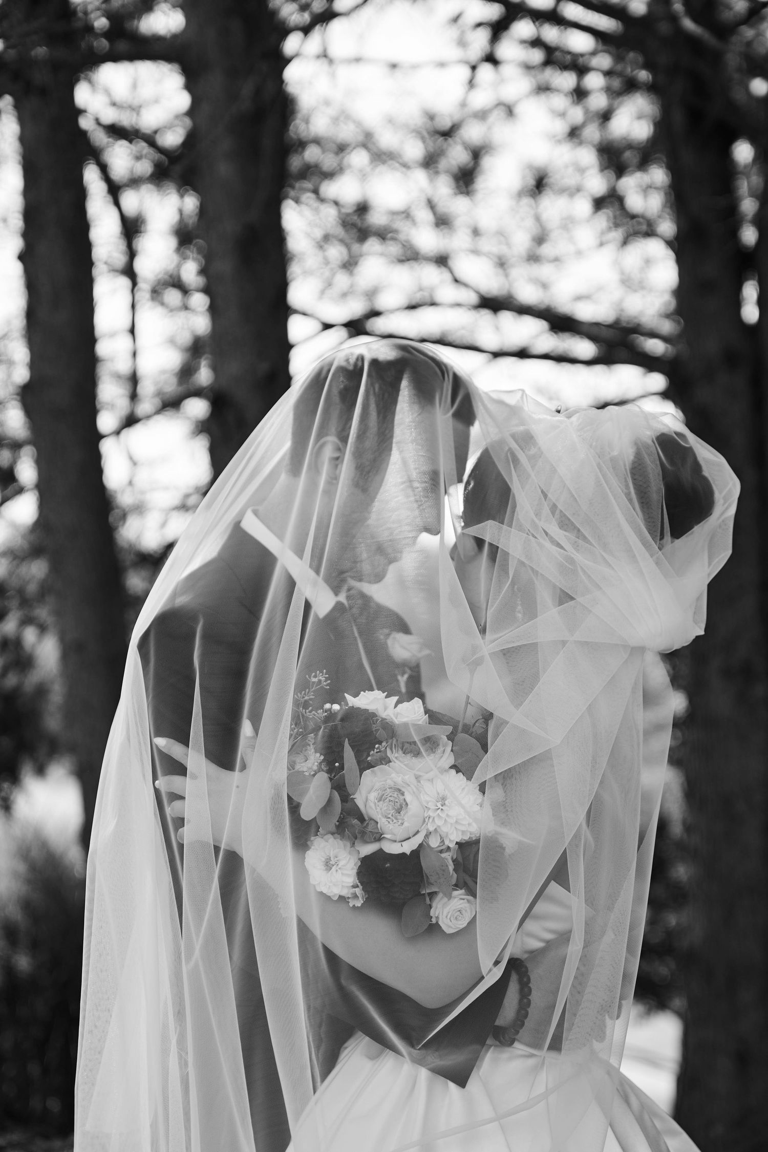 Artistic black and white shot of a bride with flowing veil holding a bouquet in a wooded setting.