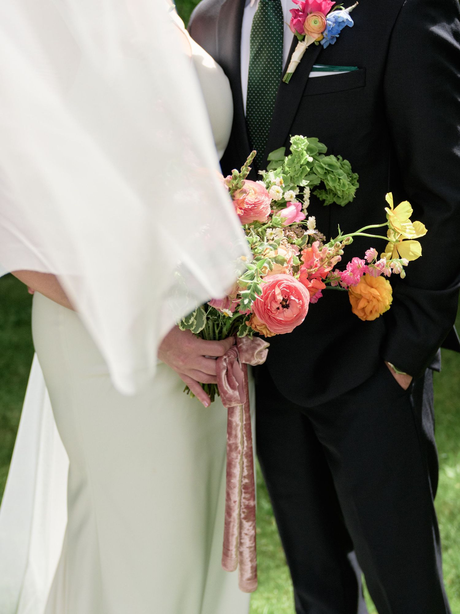 Close-up detail of a wedding bouquet featuring coral ranunculus, yellow flowers and greenery against formal attire.