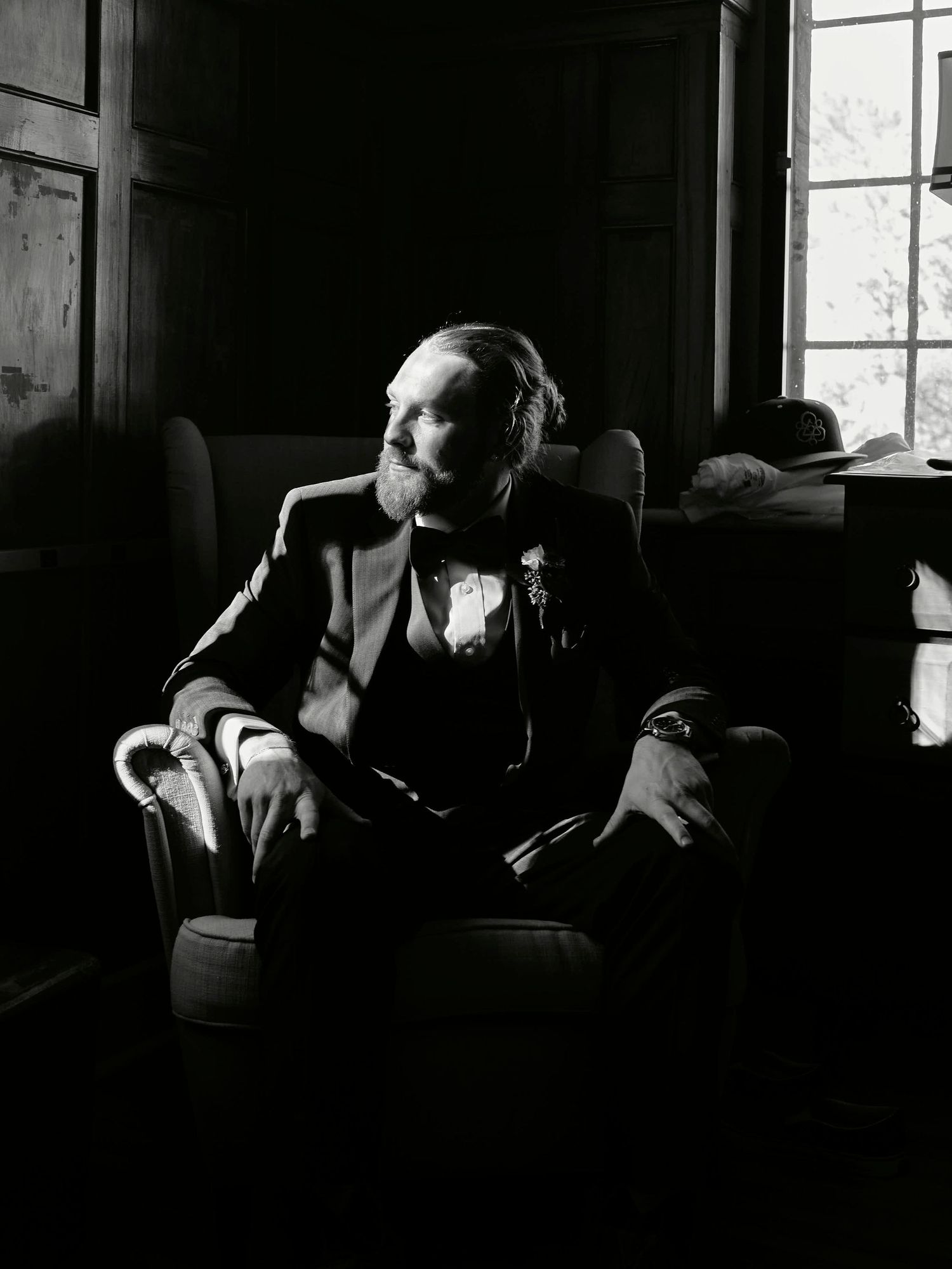 Dramatic black and white portrait of groom sitting in armchair by window.