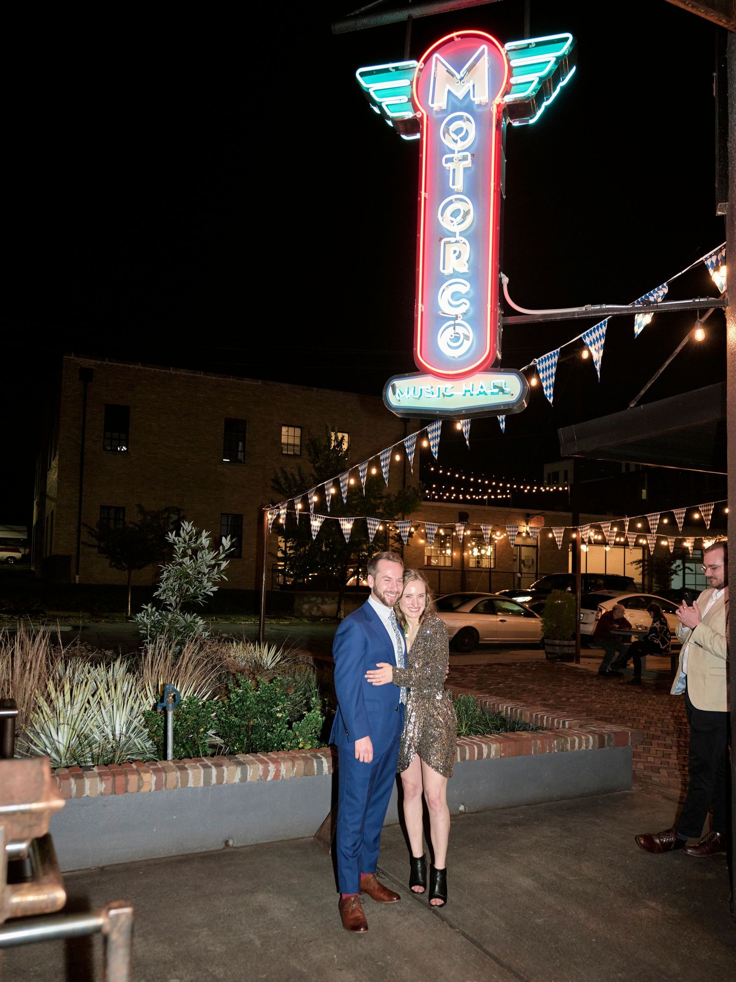 Couple poses beneath a vintage neon Motorco sign at night with string lights.
