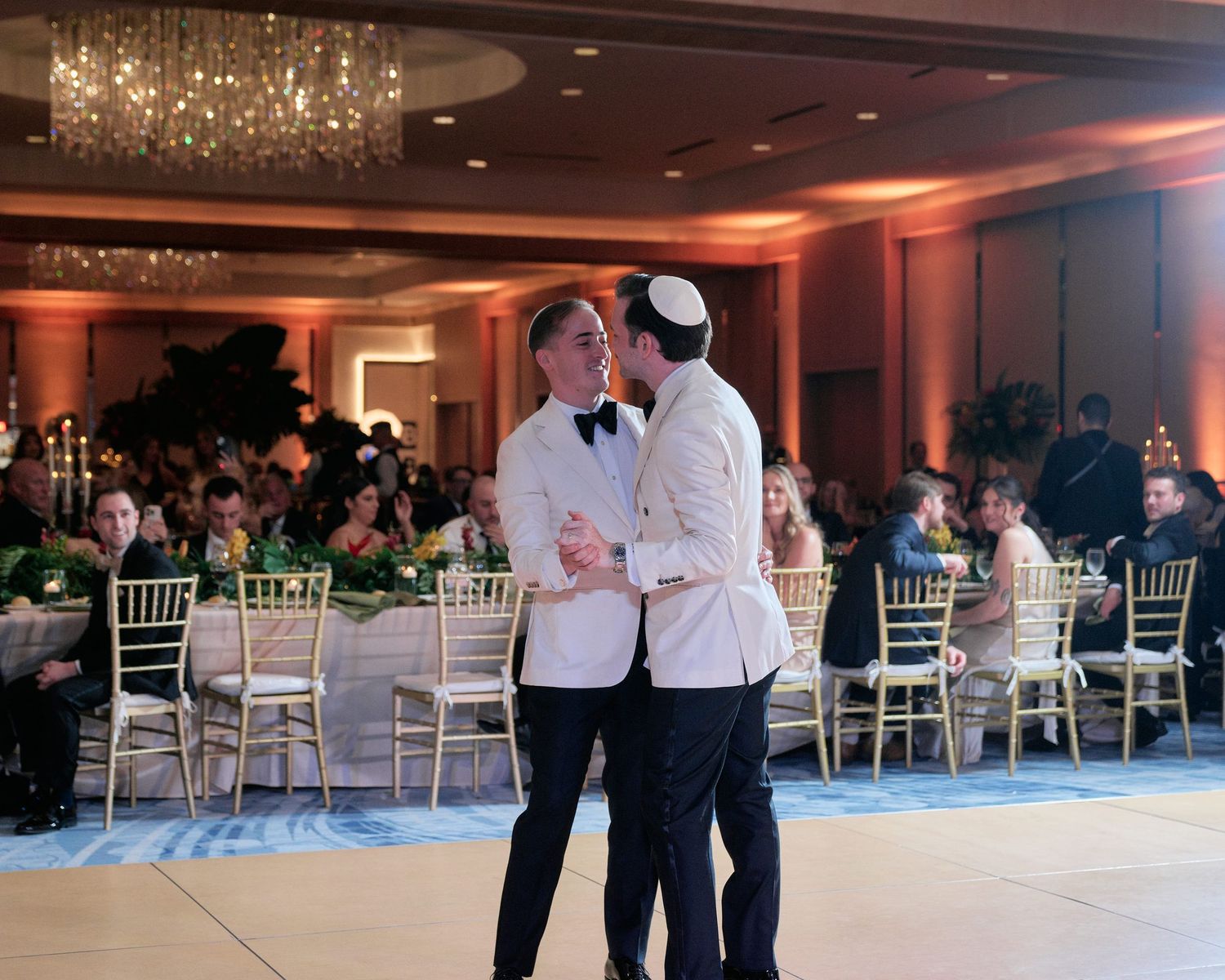 A couple shares a romantic dance at an elegant ballroom reception with chandelier lighting and white Chiavari chairs.
