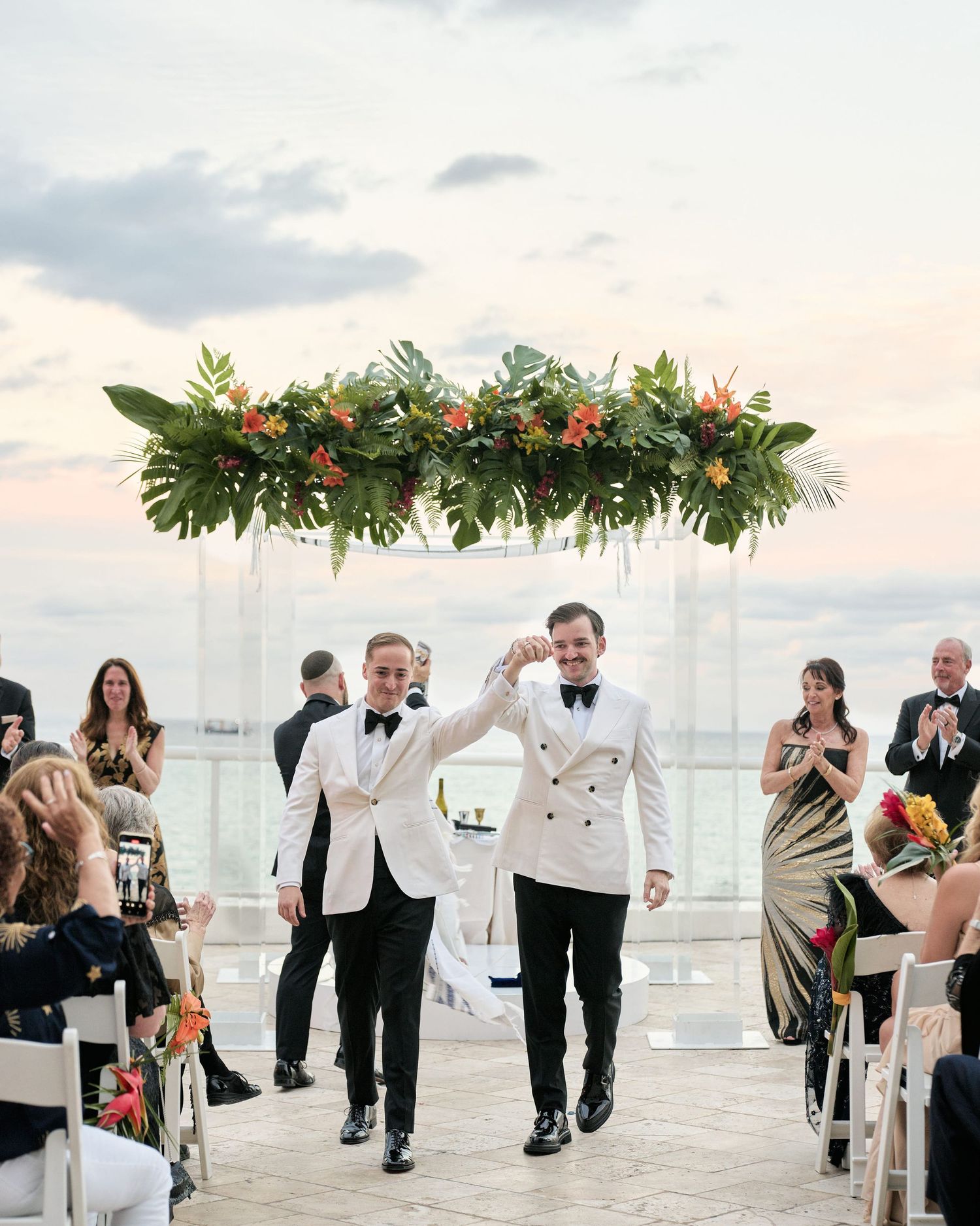 Two people walking down the aisle under a tropical floral arch during a beachside ceremony with guests watching.