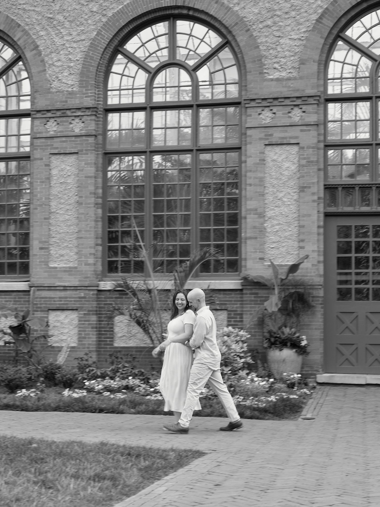 A couple walks together in black and white against an ornate brick building with arched windows.