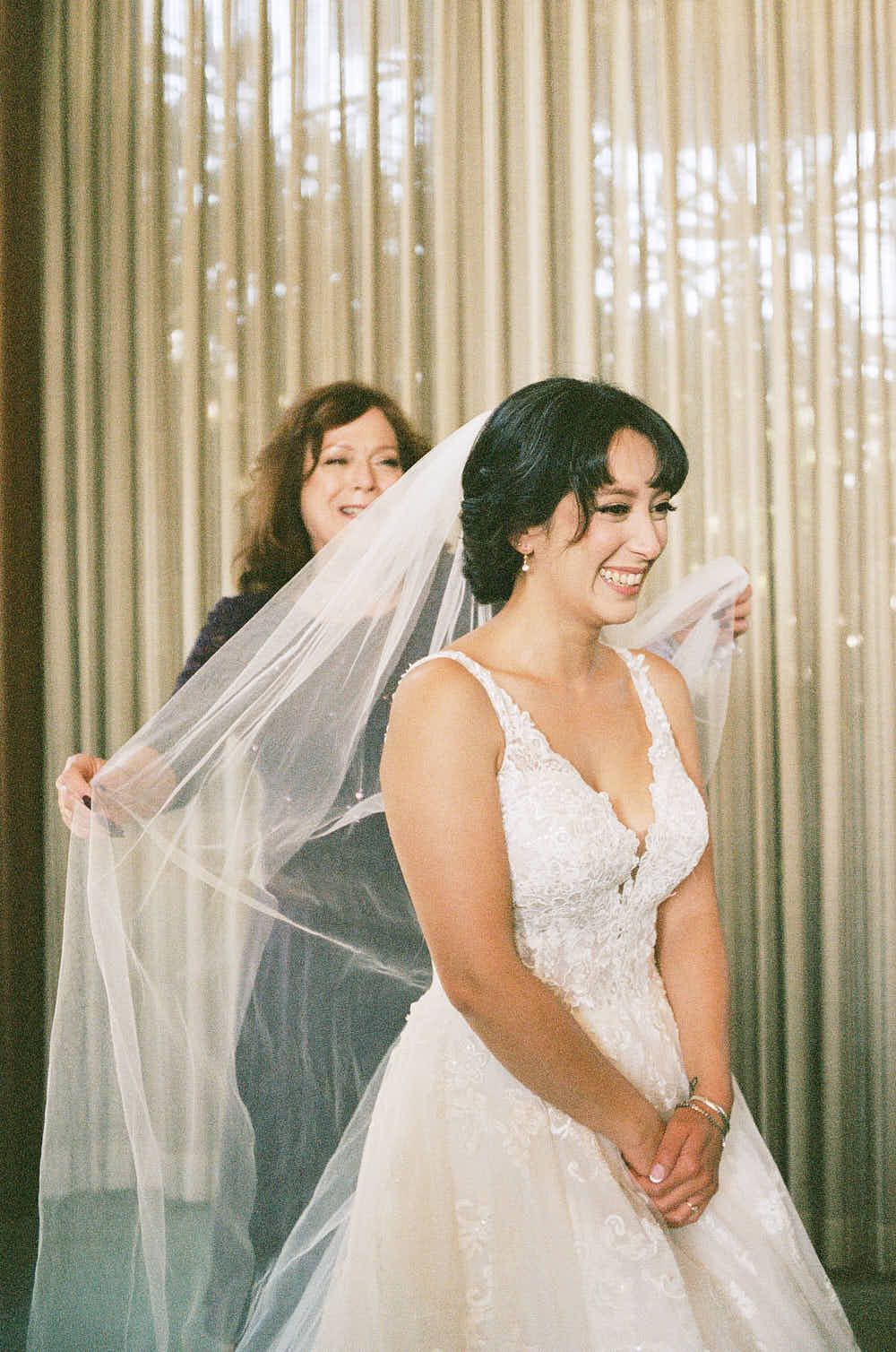 Joyful bride getting helped with veil during wedding preparations.
