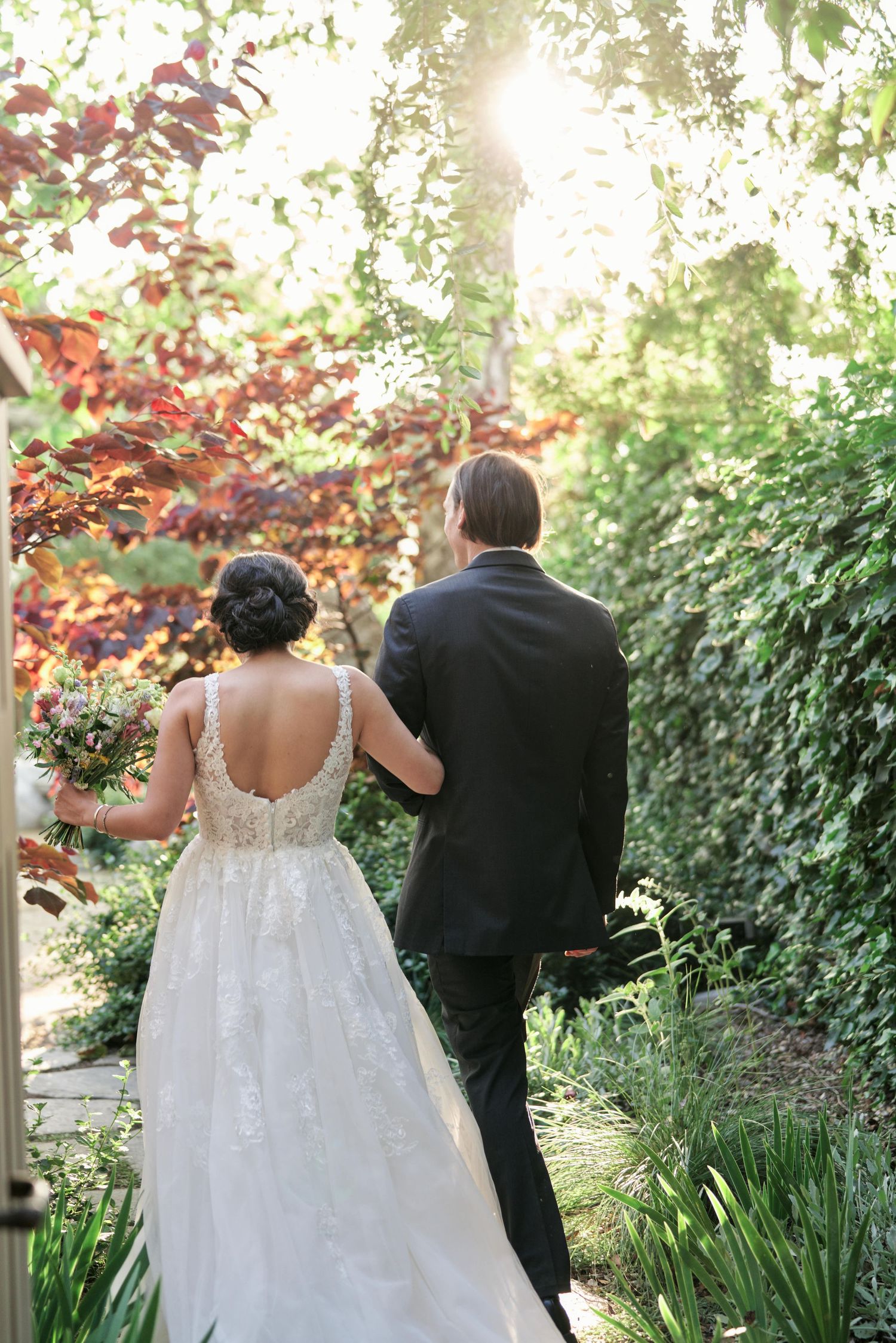 A couple walks through a sunlit garden path with autumn foliage.