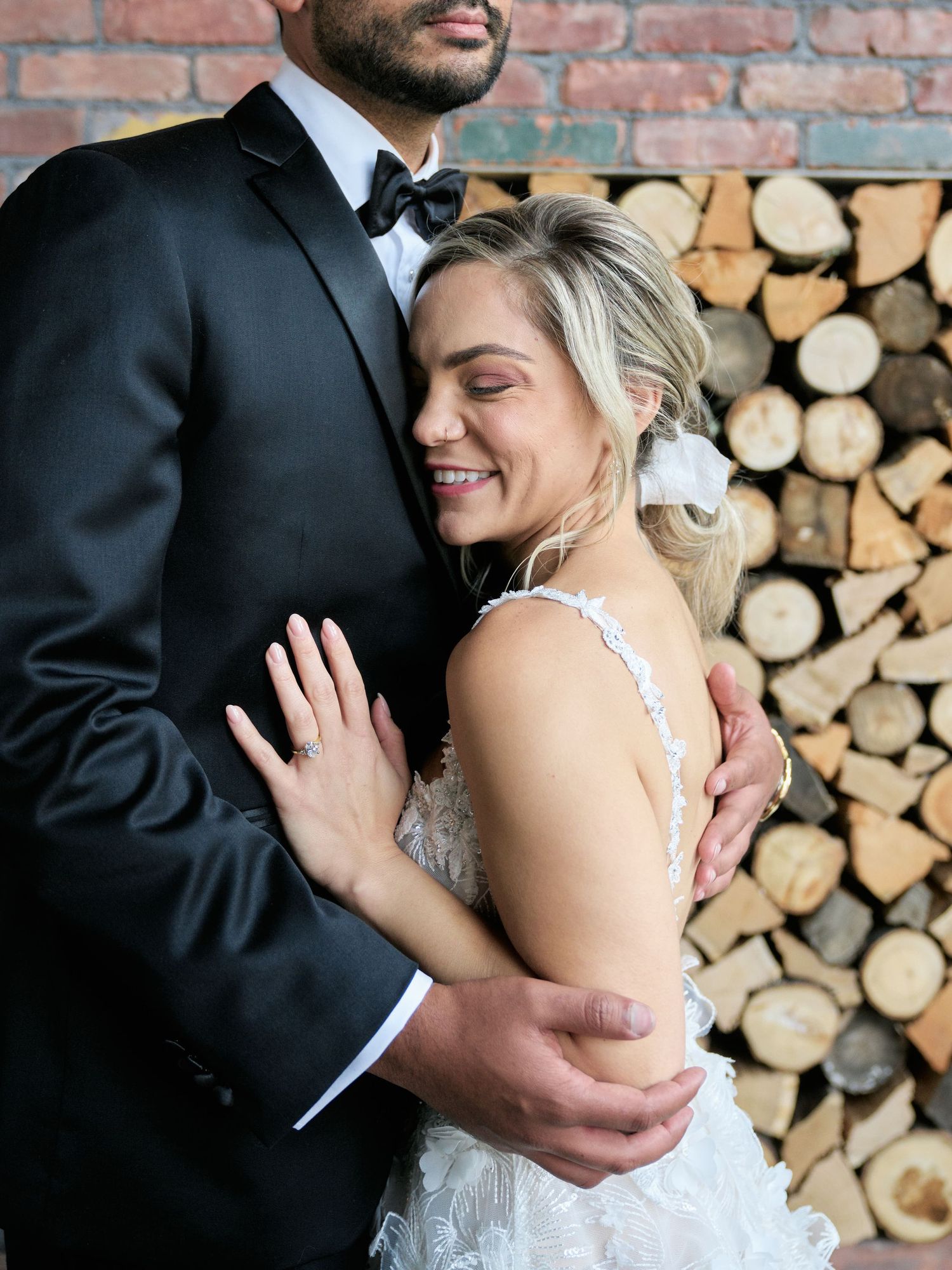 A bride and groom embrace in front of a rustic wood pile backdrop during their wedding celebration.