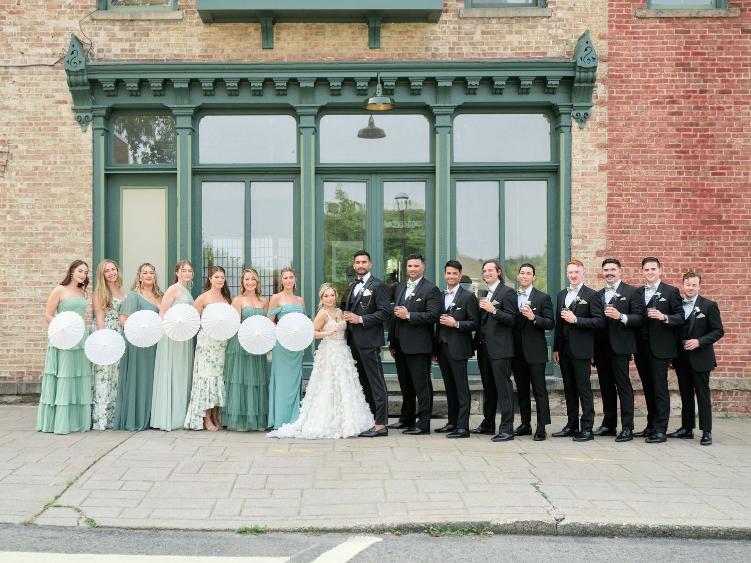 Wedding party in mint green and black formal attire lined up against vintage brick storefront with traditional architecture.