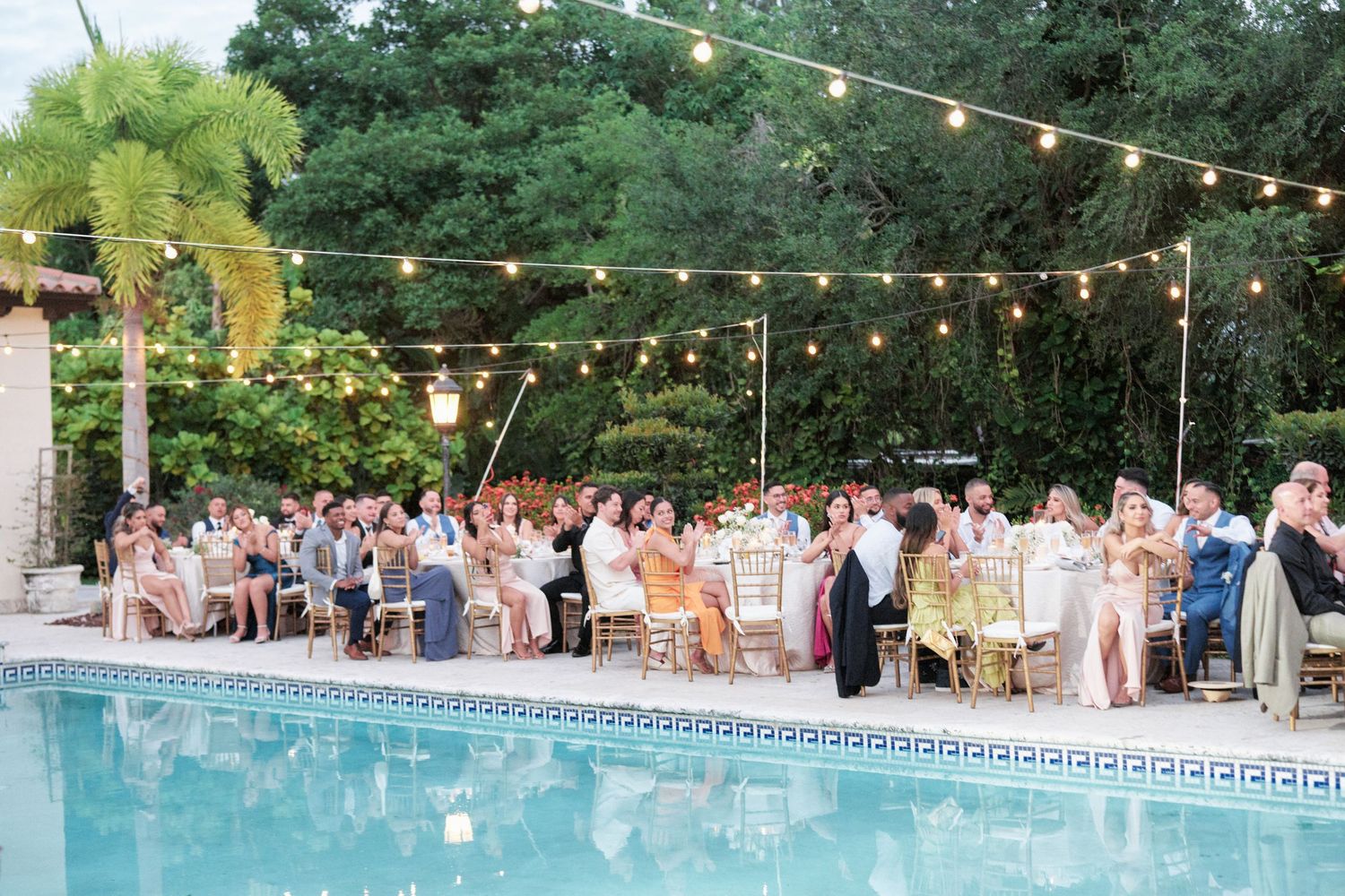 Poolside wedding reception under string lights with guests seated at round tables at dusk.