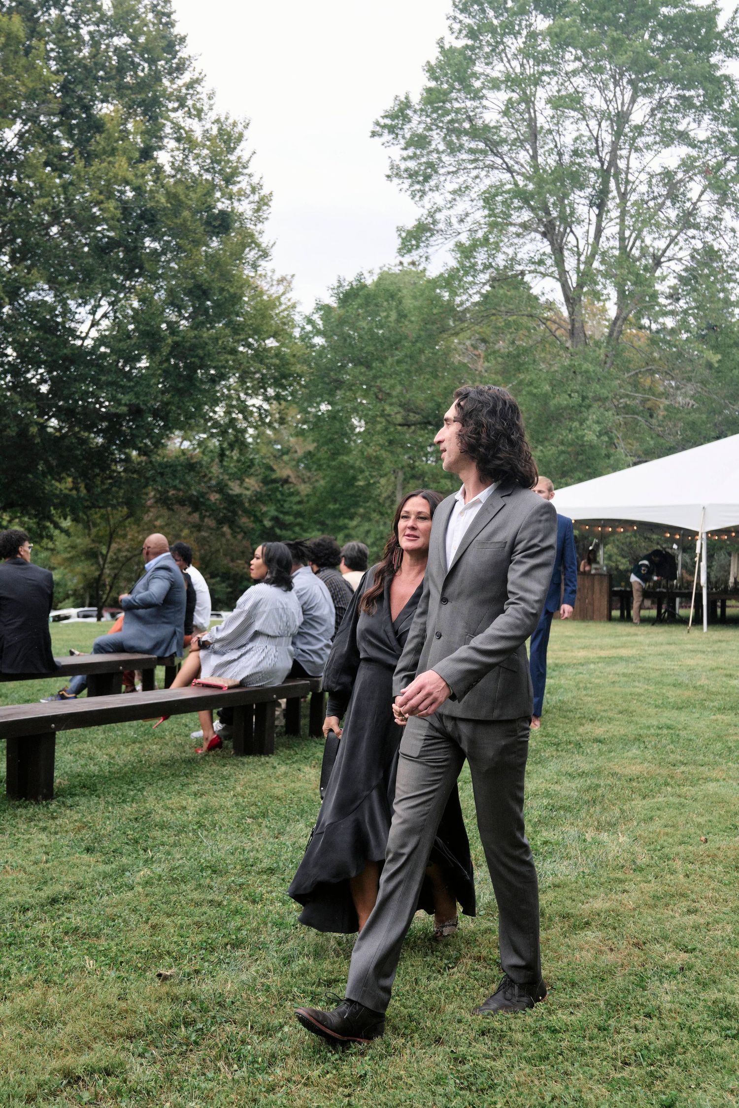 Wedding guests in formal attire stand near white tent and benches on a grassy lawn surrounded by trees.