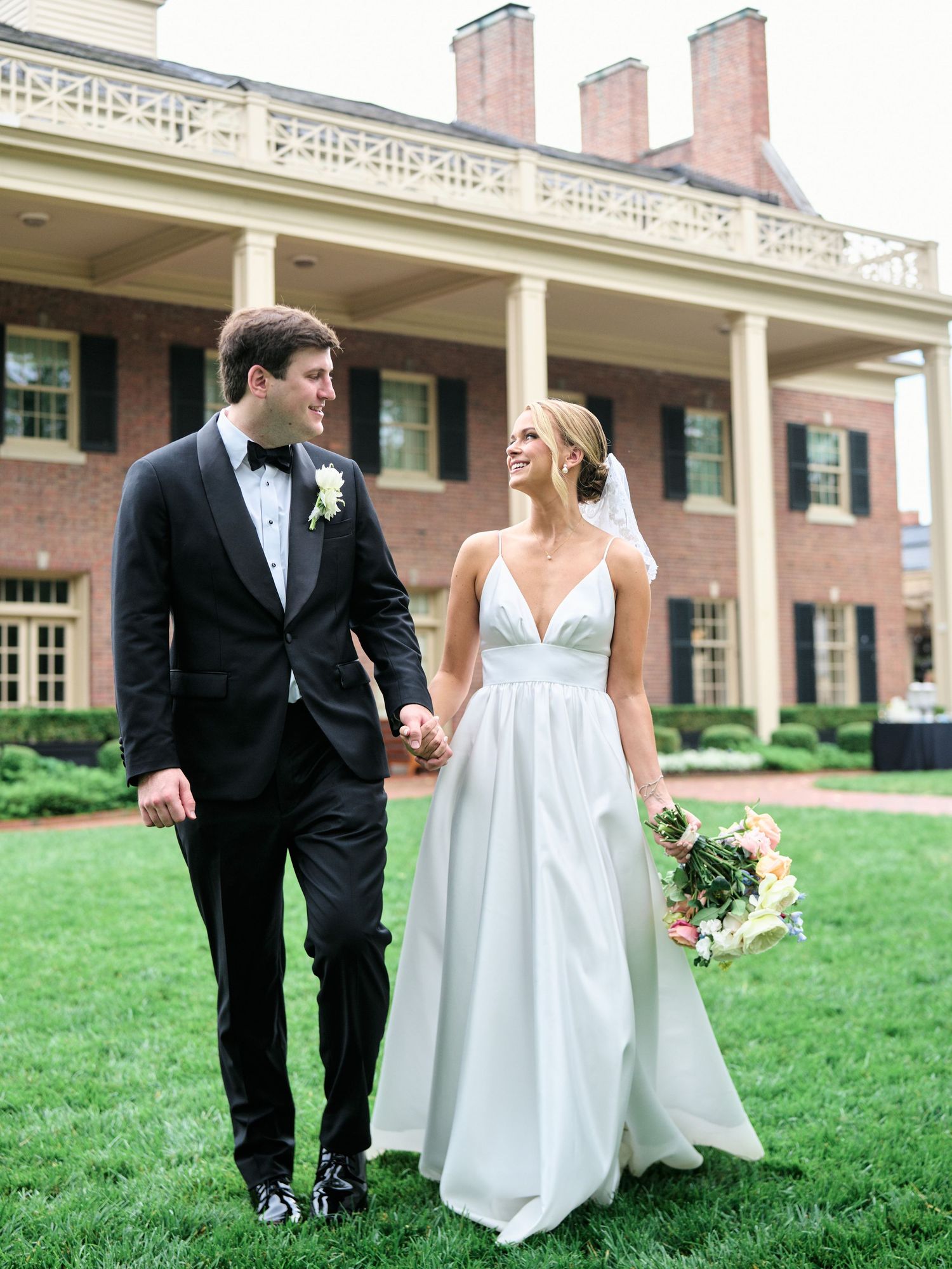 Couple holding hands while walking on grass in front of a historic brick building with columns.