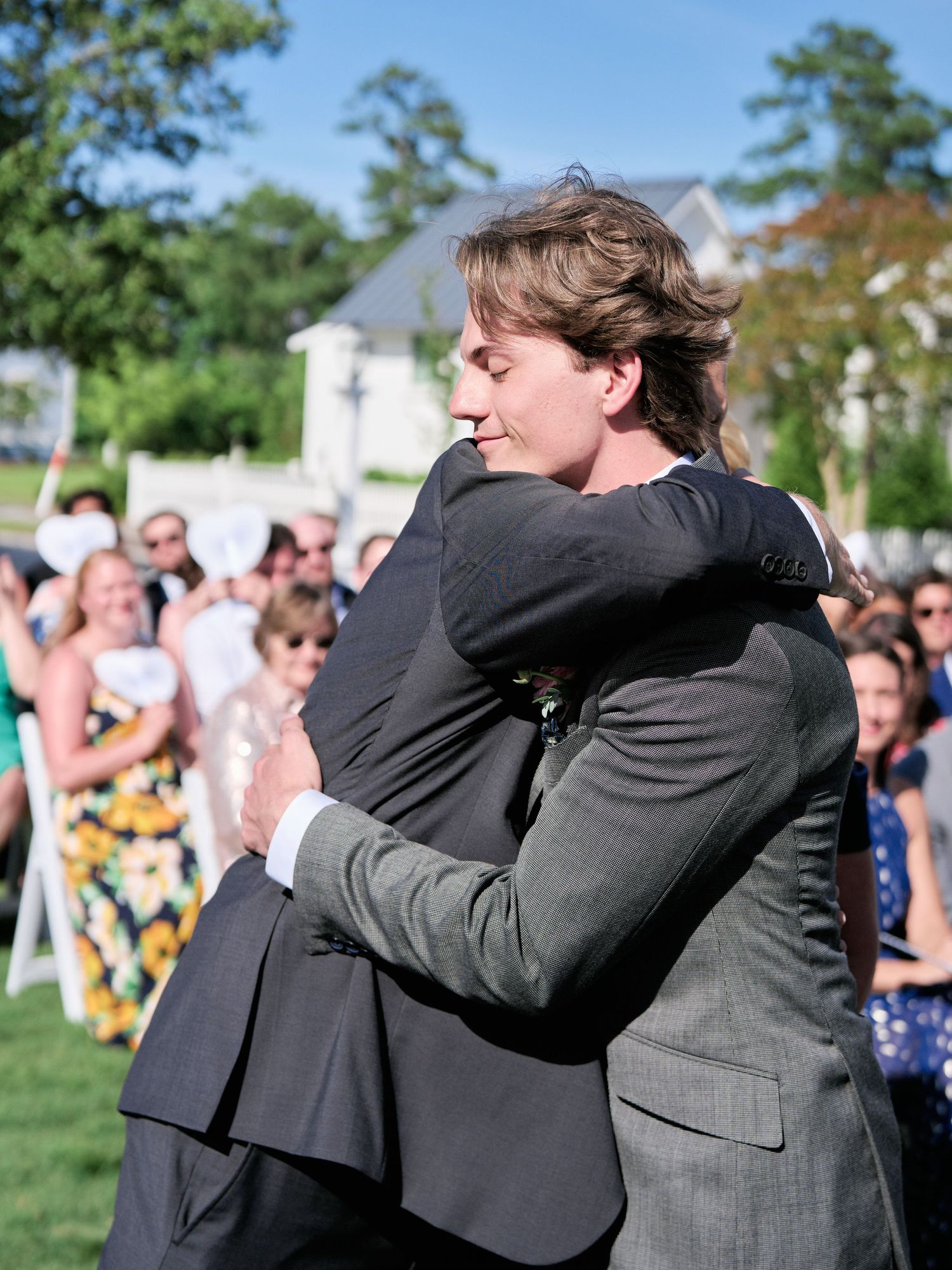 Two people share an emotional embrace outdoors at an event with a crowd gathered behind them.