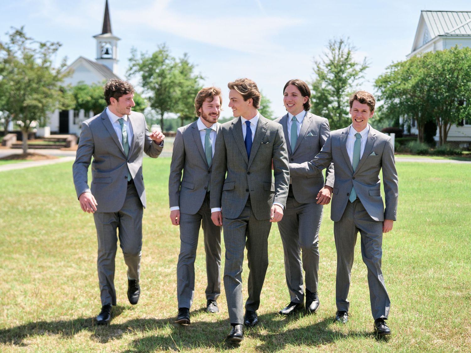 Groomsmen in matching gray suits walking and laughing together on a lawn with a white church steeple in background.