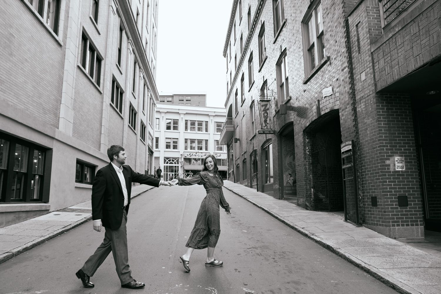 A black and white photo of a couple dancing playfully on an empty urban street.