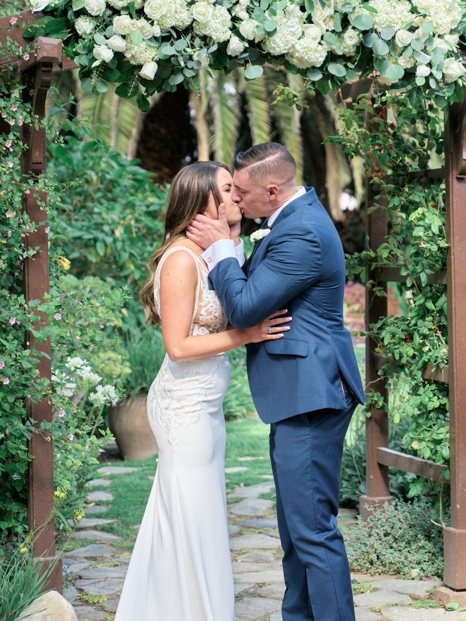 Wedding couple kissing under floral arch with white roses and greenery.