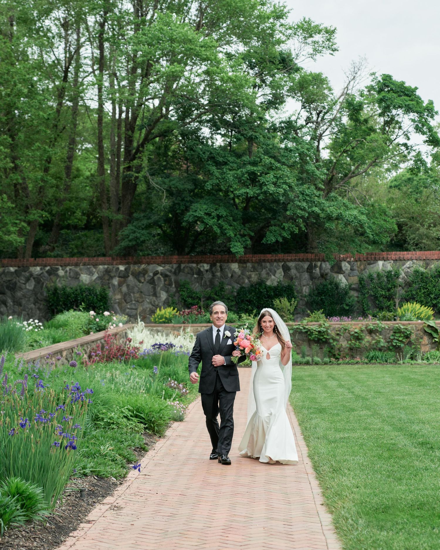 A couple walks along a brick pathway bordered by lush gardens and a stone wall in an outdoor setting.
